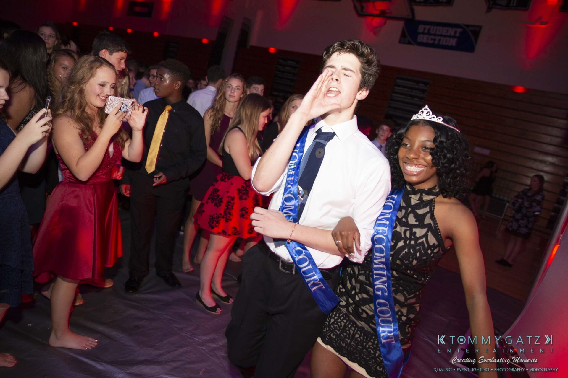 SR-Homecoming-102718-Carl-Low-Res-49 - TOMMY GATZ ENTERTAINMENT Students wearing sashes at a homecoming event, one holding a microphone, surrounded by classmates on the dance floor.