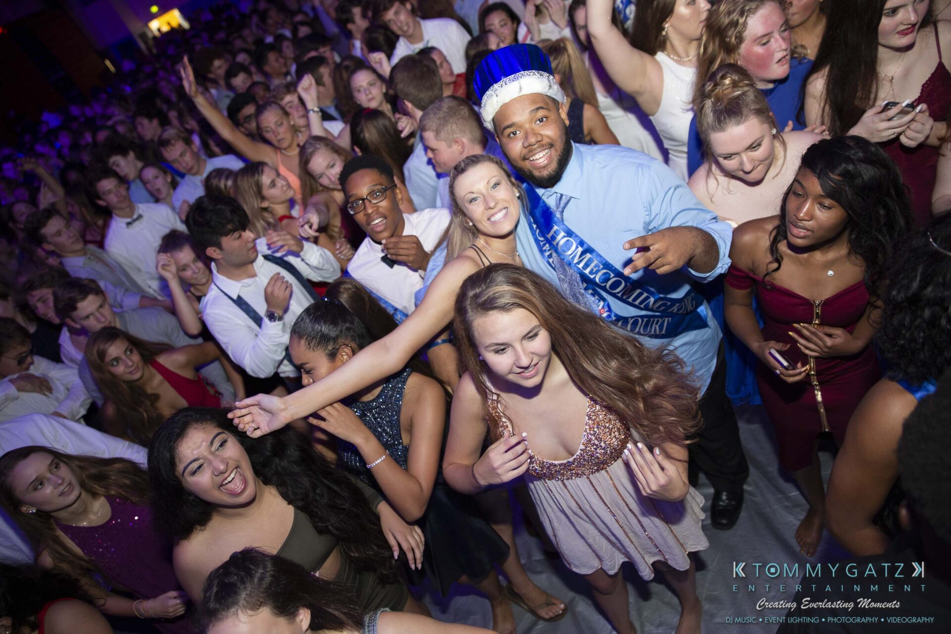 SR-Homecoming-102718-Carl-Low-Res-55 - TOMMY GATZ ENTERTAINMENT A group of people dressed in formal attire smile and dance at a crowded indoor event with festive lighting.
