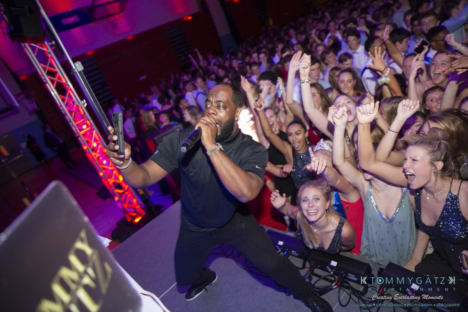 SR-Homecoming-102718-Carl-Low-Res-56 - TOMMY GATZ ENTERTAINMENT DJ performing on stage with a microphone, as a lively crowd dances and cheers in a colorful, well-lit venue.