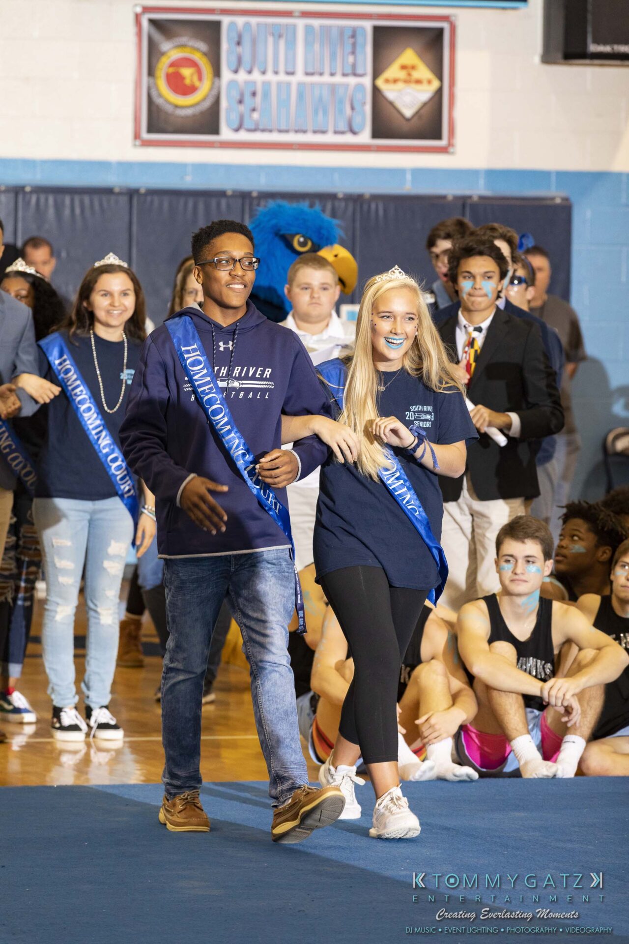 SR-Pep-Rally-102618-Carl-Low-Res-10 - TOMMY GATZ ENTERTAINMENT Two students wearing sashes walk on a blue carpet at an indoor event, surrounded by others.