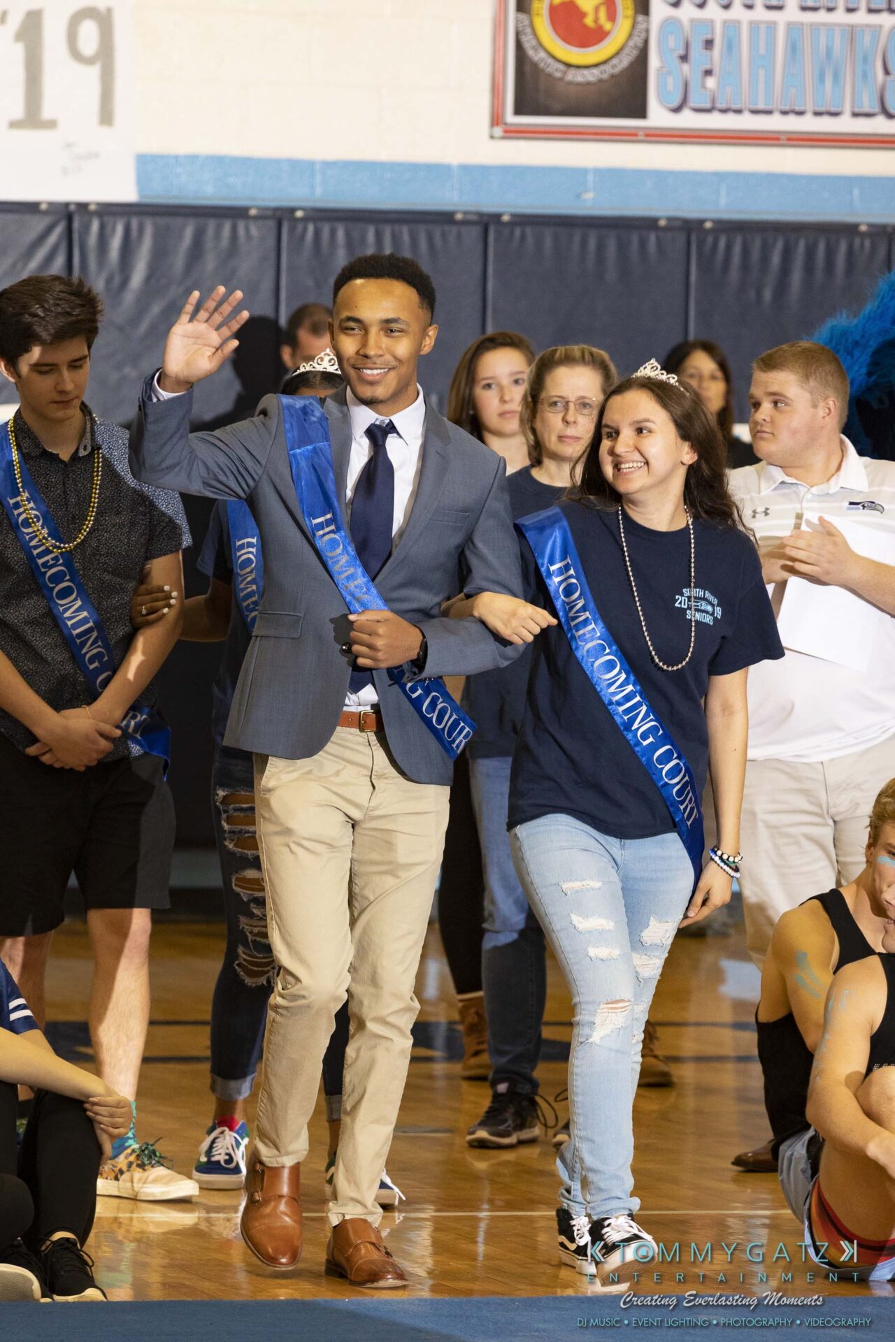 SR-Pep-Rally-102618-Carl-Low-Res-11 - TOMMY GATZ ENTERTAINMENT Two people in blue sashes walk arm in arm at a school event, with onlookers in the background.