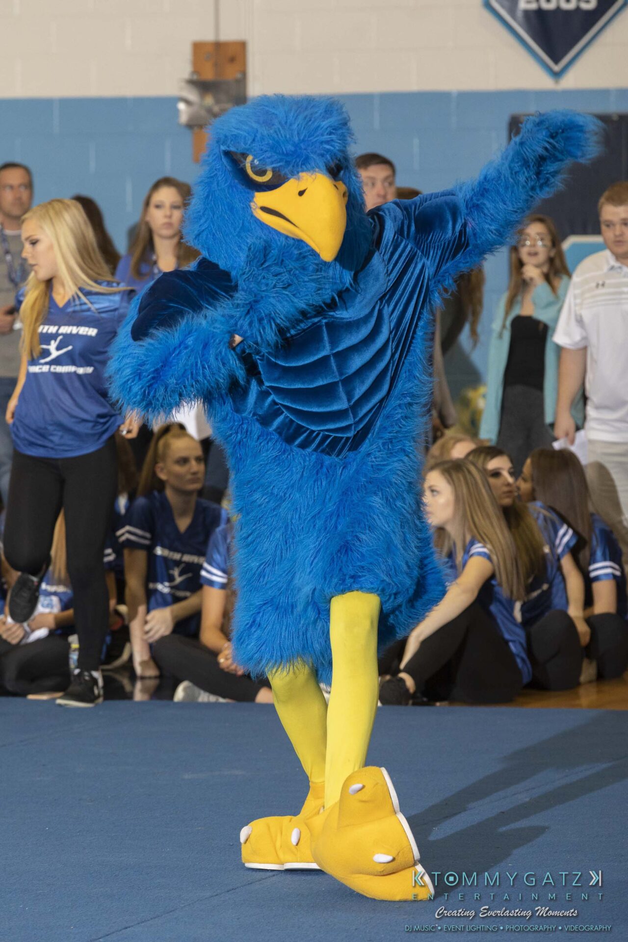 SR-Pep-Rally-102618-Carl-Low-Res-2 - TOMMY GATZ ENTERTAINMENT A person in a blue and yellow bird mascot costume poses on a gymnasium floor. A crowd of people stands in the background.
