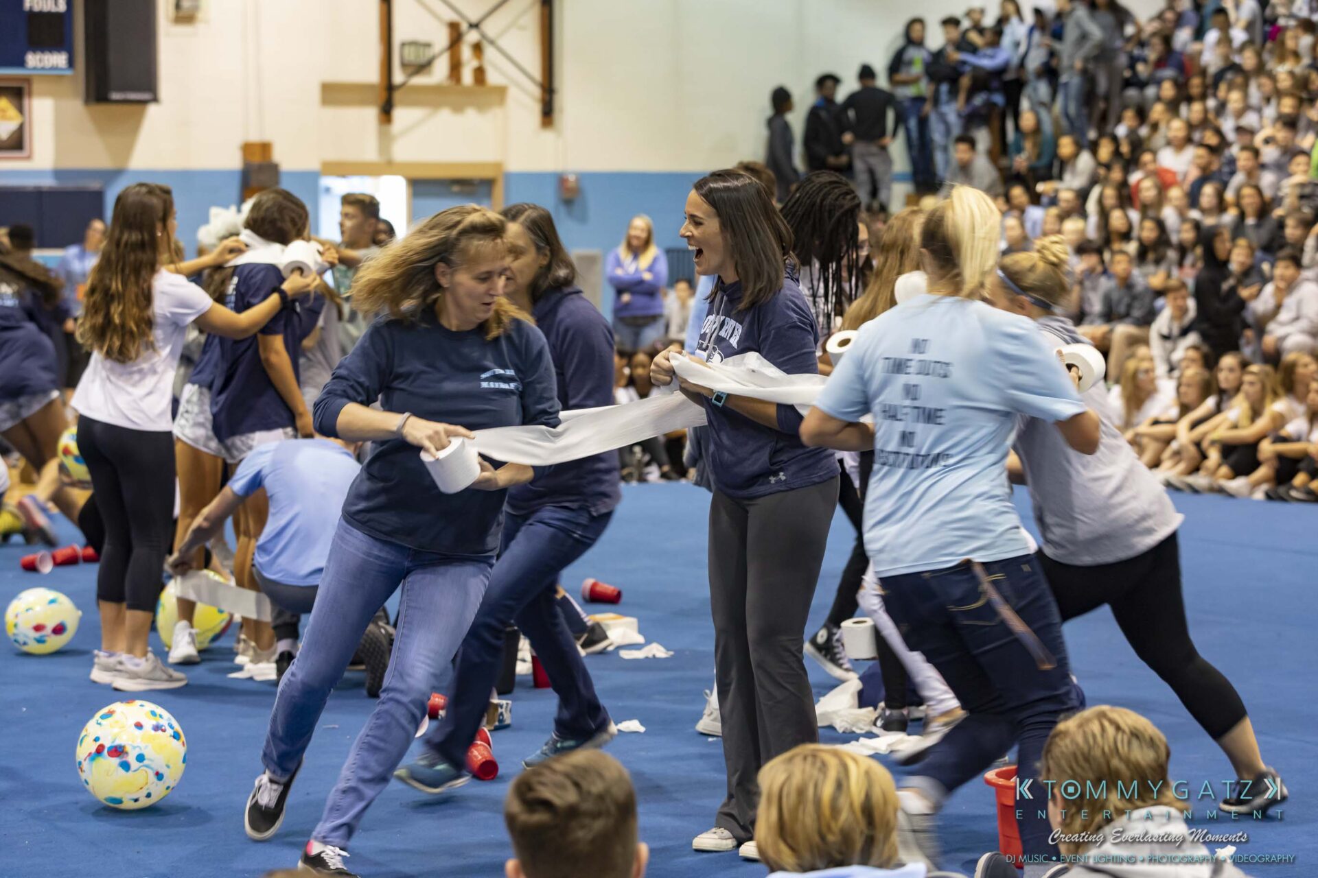 SR-Pep-Rally-102618-Carl-Low-Res-43 - TOMMY GATZ ENTERTAINMENT People participating in a lively indoor game involving toilet paper in a gymnasium, with a seated audience watching.