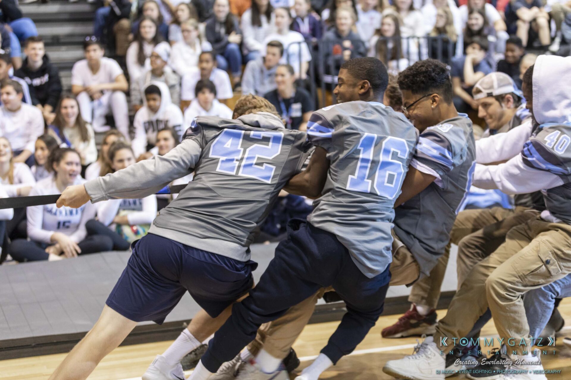 SR-Pep-Rally-102618-Carl-Low-Res-55 - TOMMY GATZ ENTERTAINMENT Three young men in numbered jerseys participate in a tug-of-war, surrounded by seated spectators in a gymnasium.