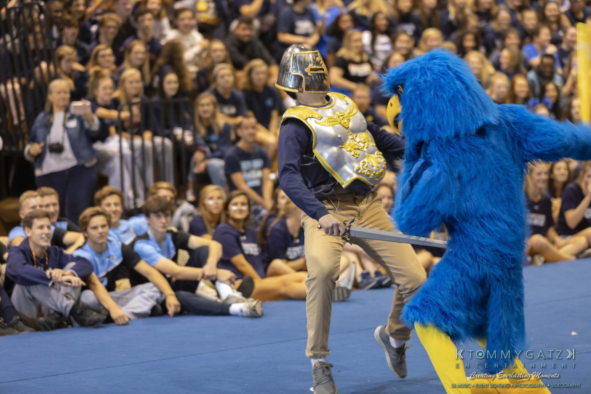 SR-Pep-Rally-102618-Carl-Low-Res-64 - TOMMY GATZ ENTERTAINMENT A person in a knight costume playfully interacts with a person in a blue bird costume on a gym floor surrounded by a seated crowd.