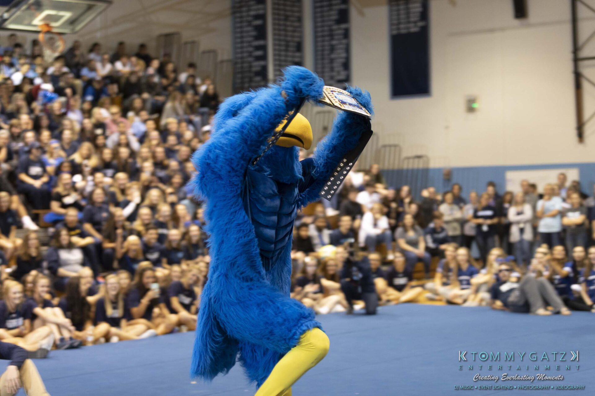 SR-Pep-Rally-102618-Carl-Low-Res-65 - TOMMY GATZ ENTERTAINMENT Blue bird mascot running in a gymnasium with a cheering crowd in the background.