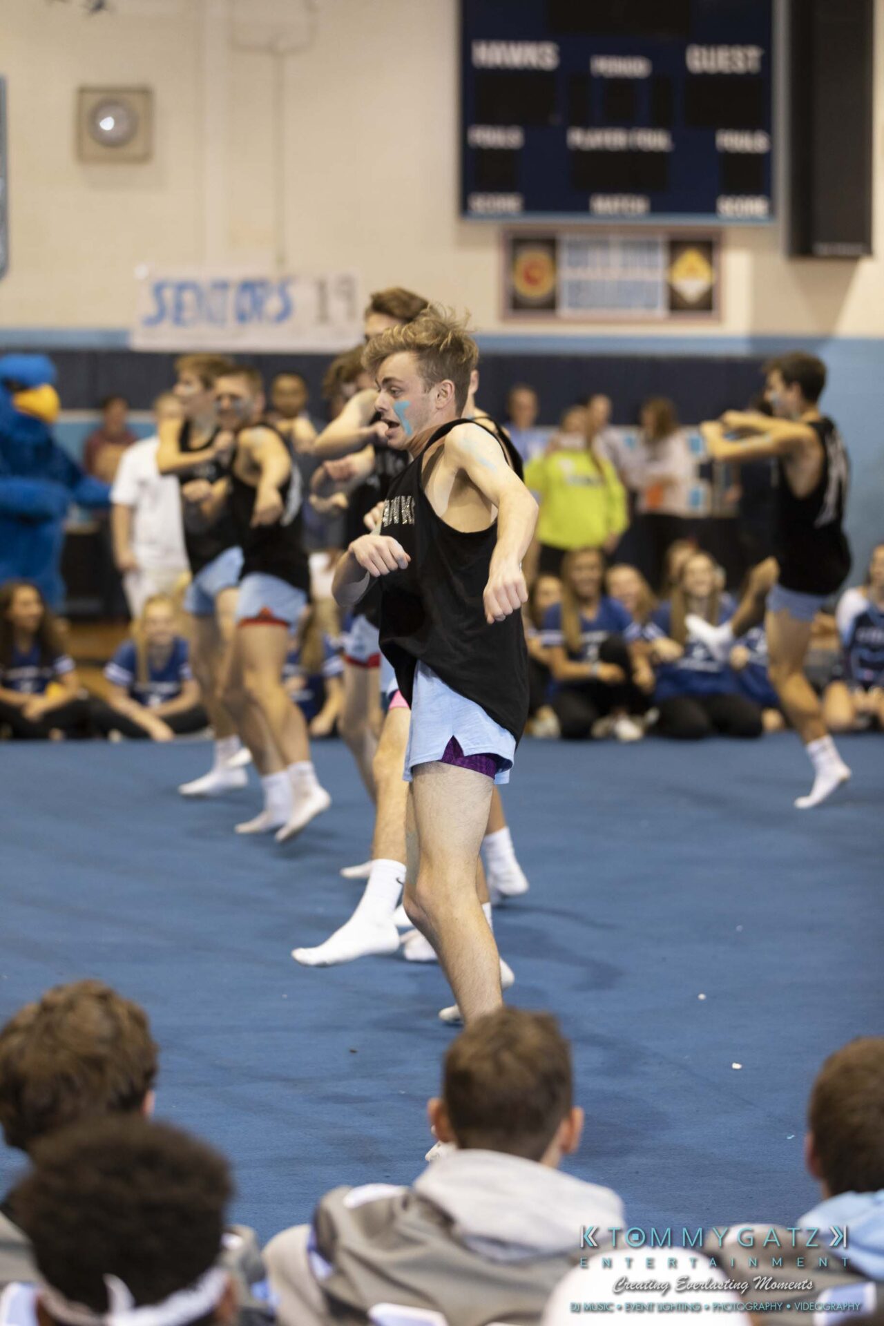 SR-Pep-Rally-102618-Carl-Low-Res-66 - TOMMY GATZ ENTERTAINMENT A male cheerleader performs a high kick during a routine at a school gym. Students and cheerleaders watch from the background.