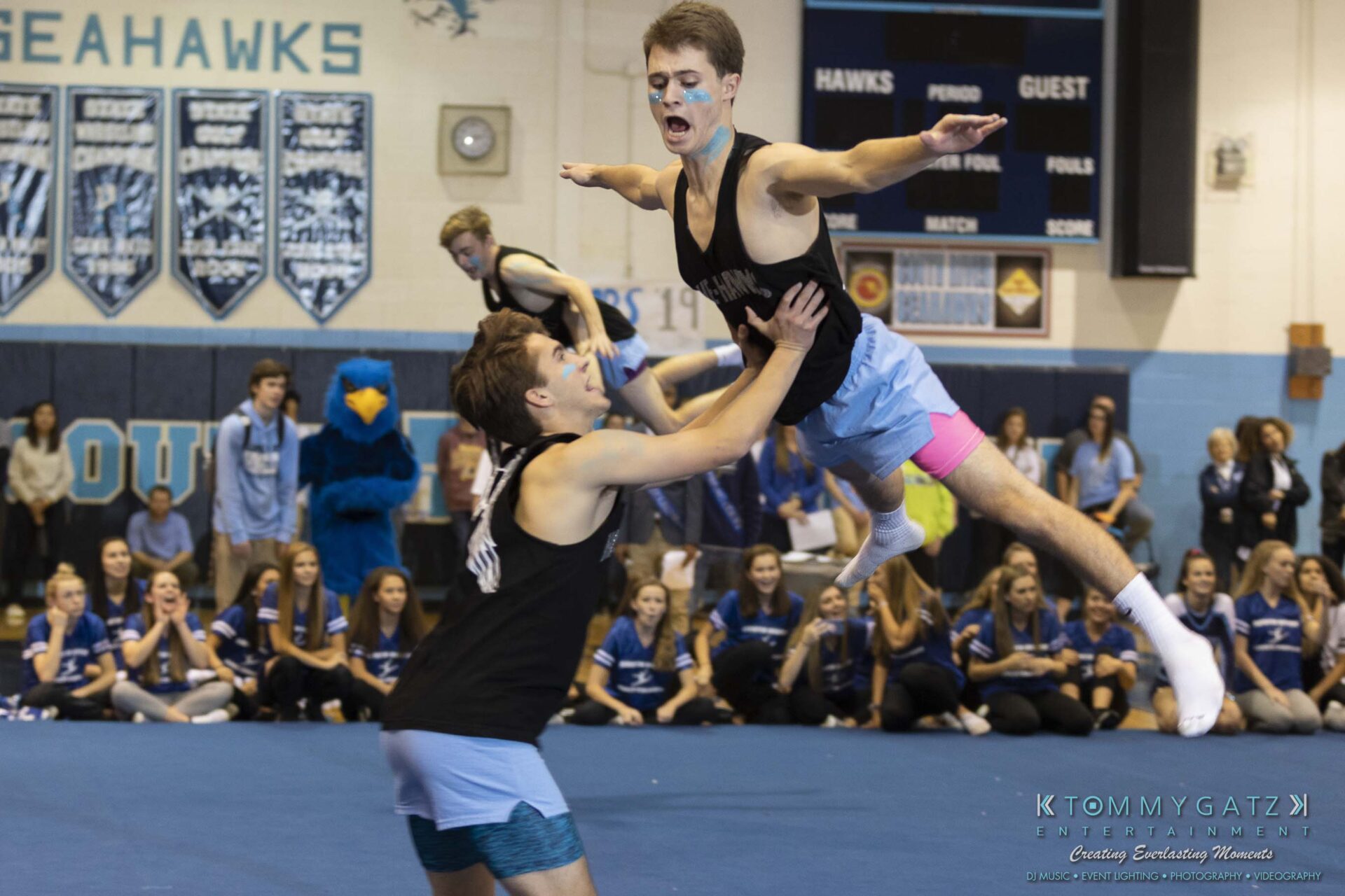 SR-Pep-Rally-102618-Carl-Low-Res-67 - TOMMY GATZ ENTERTAINMENT Two men performing a cheerleading stunt in a gym. One is being lifted horizontally by the other. A crowd sits in the background watching.