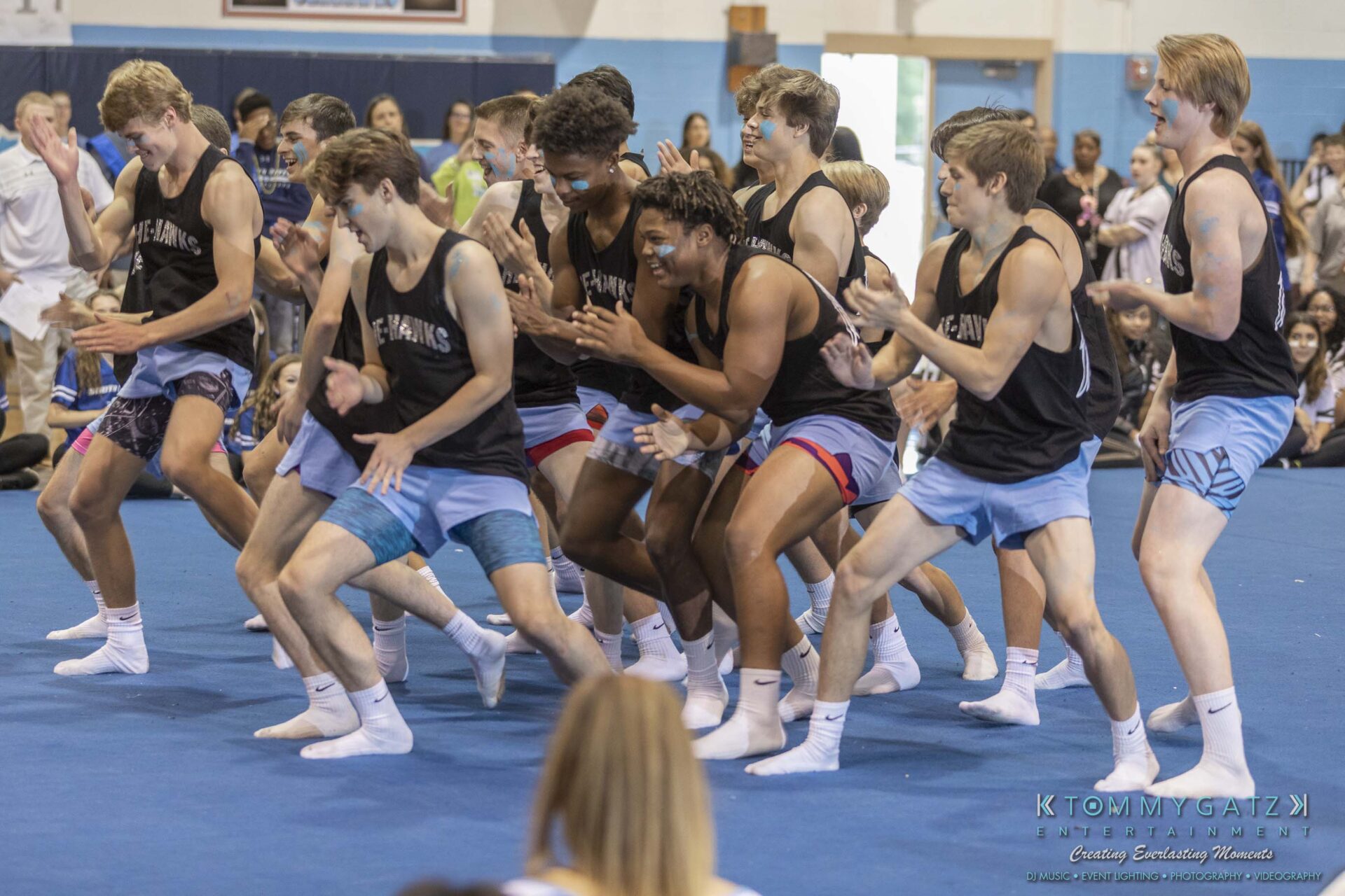 SR-Pep-Rally-102618-Carl-Low-Res-68 - TOMMY GATZ ENTERTAINMENT A group of young men in athletic wear perform a synchronized dance routine in a gymnasium, with an audience watching in the background.