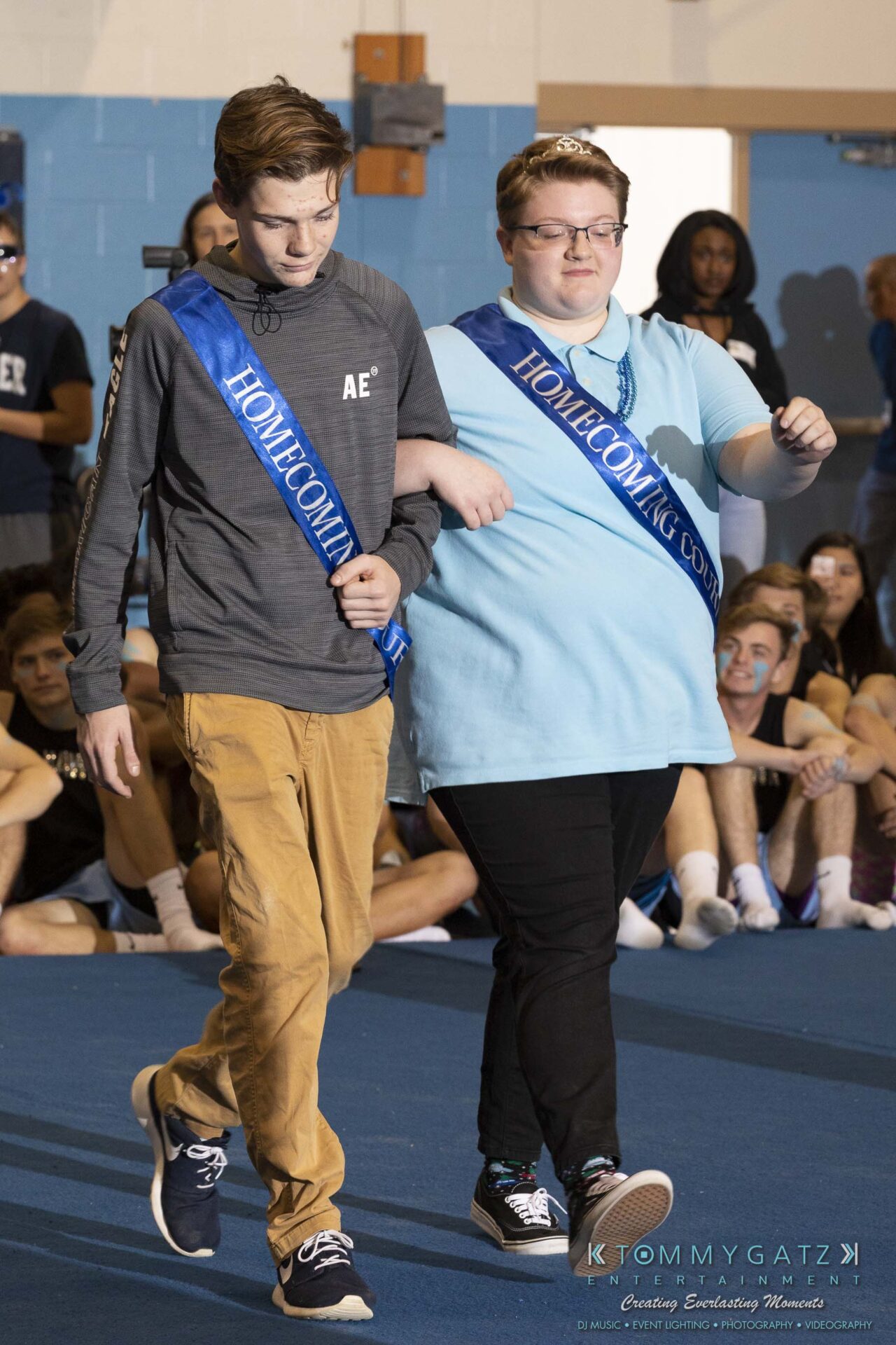 SR-Pep-Rally-102618-Carl-Low-Res-7 - TOMMY GATZ ENTERTAINMENT Two individuals wearing blue "Homecoming Court" sashes walk together in a gymnasium, with a seated crowd in the background.
