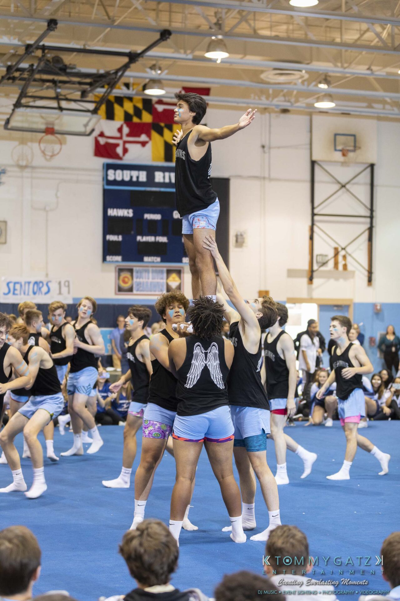 SR-Pep-Rally-102618-Carl-Low-Res-70 - TOMMY GATZ ENTERTAINMENT Cheerleading team performing a group stunt in a gymnasium, with one member lifted in the air while others support from below. Banners and a scoreboard are visible in the background.