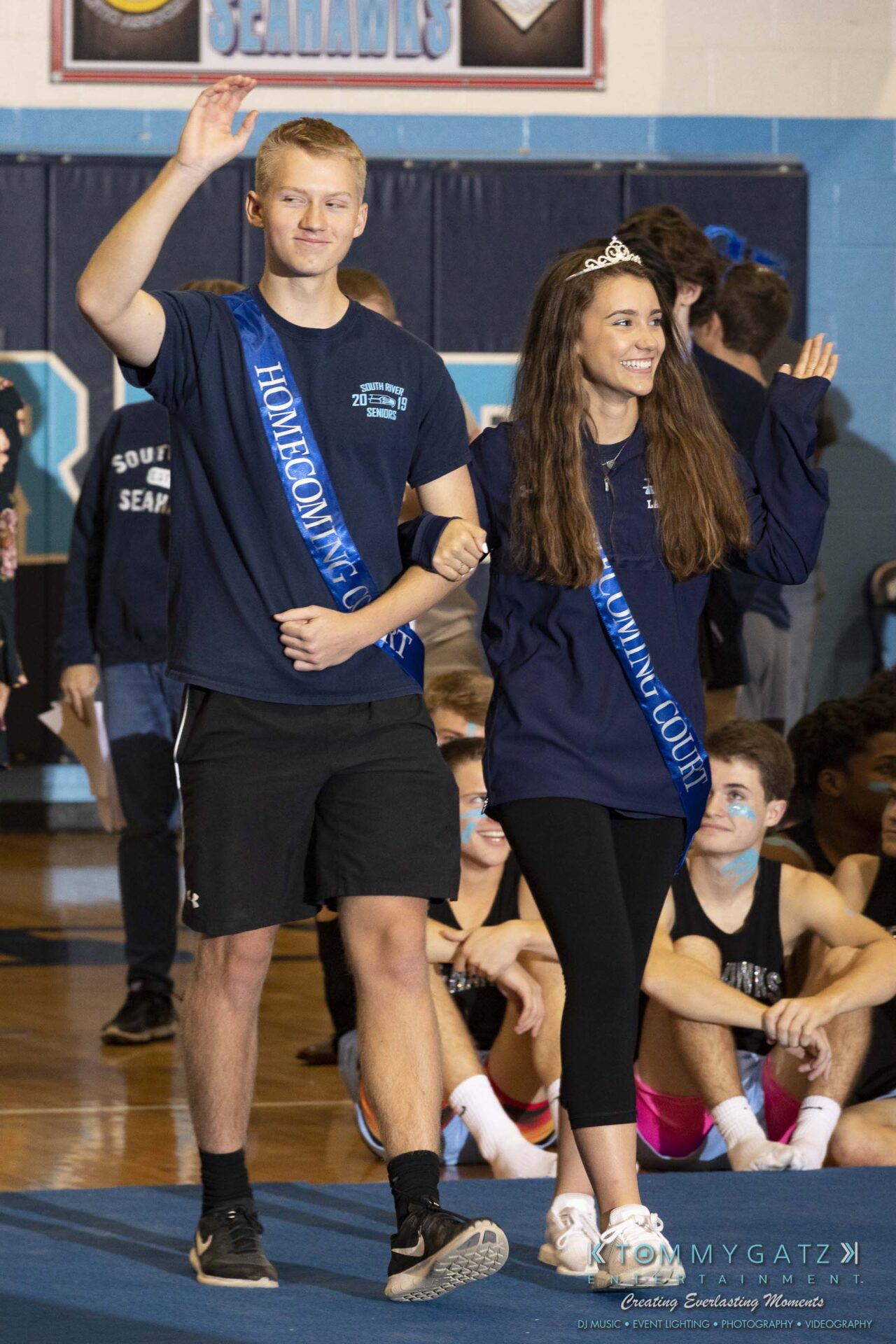 SR-Pep-Rally-102618-Carl-Low-Res-8 - TOMMY GATZ ENTERTAINMENT A young man and woman wearing homecoming sashes and casual attire walk arm in arm in a gym, with the man waving.