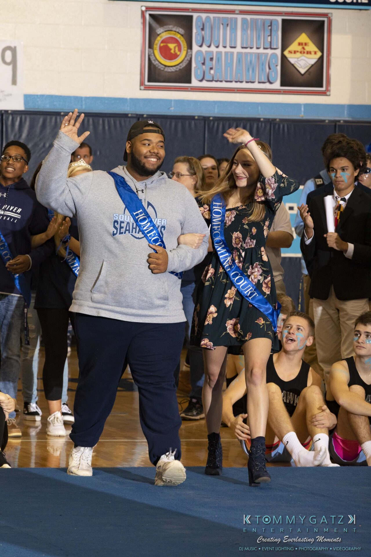 SR-Pep-Rally-102618-Carl-Low-Res-9 - TOMMY GATZ ENTERTAINMENT Two people wearing "Homecoming Court" sashes walk in a gymnasium. A group of seated and standing individuals watches in the background.