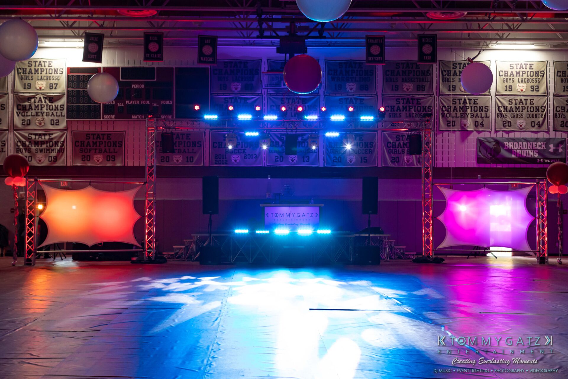 092819-Broadneck-Homecoming-Low-Res-WM-1 - TOMMY GATZ ENTERTAINMENT Indoor venue with colorful stage lights, banners on walls, and a DJ booth on a sports court. Various lighting equipment and speakers are set up around the stage area.