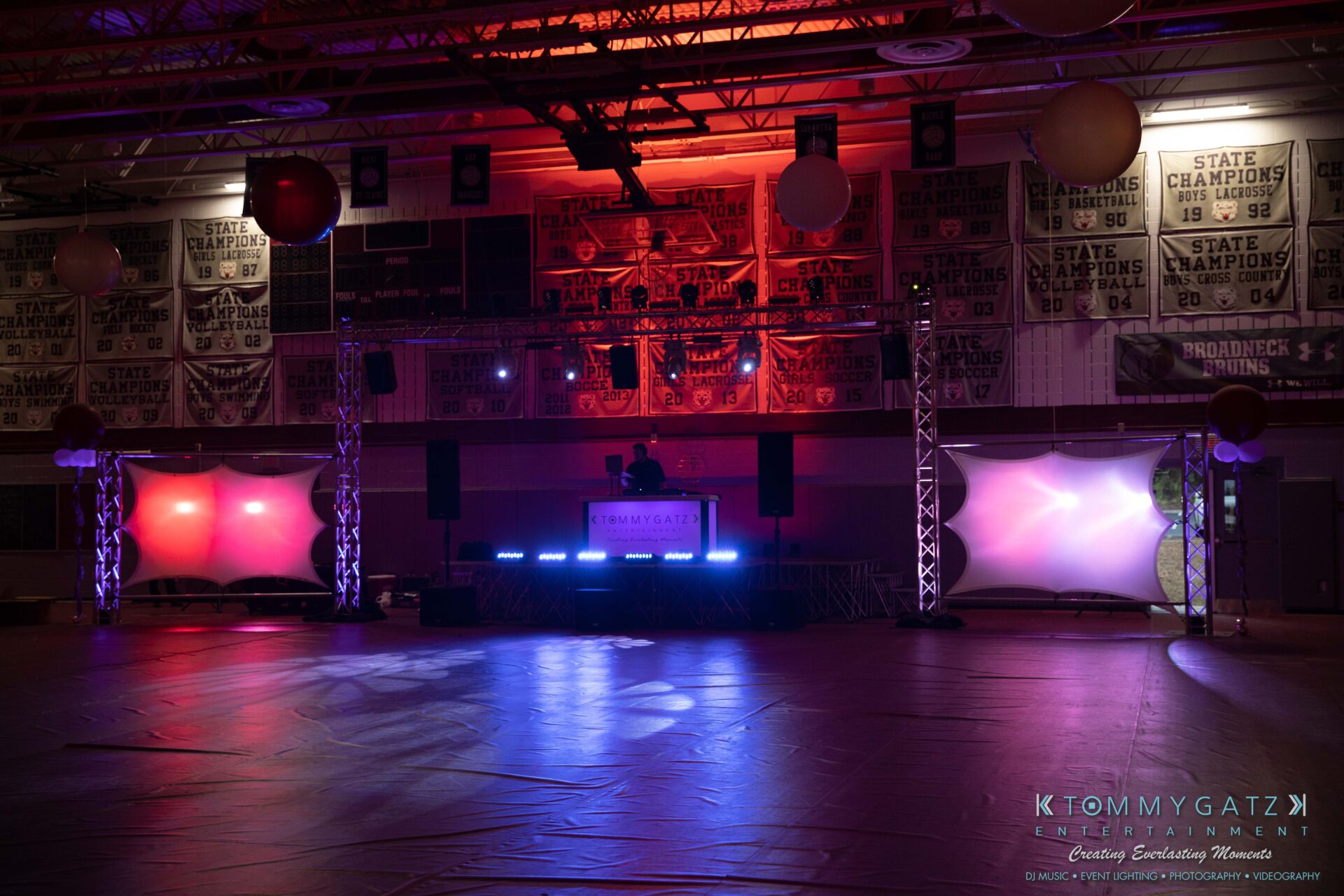 092819-Broadneck-Homecoming-Low-Res-WM-5-2 - TOMMY GATZ ENTERTAINMENT A dimly lit gymnasium decorated for an event, featuring balloons and stage lighting. A DJ booth is set up on an elevated platform under red and blue lights.