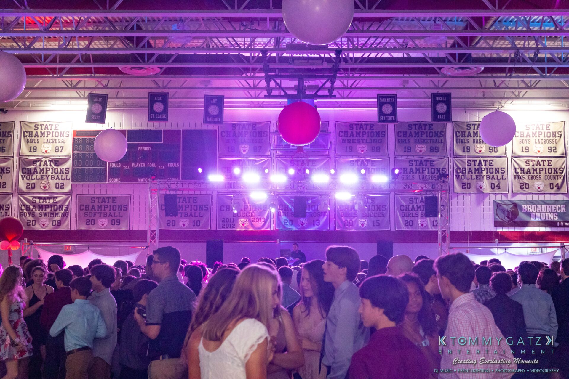 092819-Broadneck-Homecoming-Low-Res-WM-63 - TOMMY GATZ ENTERTAINMENT A crowd of people dancing in a gymnasium adorned with championship banners, colorful balloons, and bright stage lights.