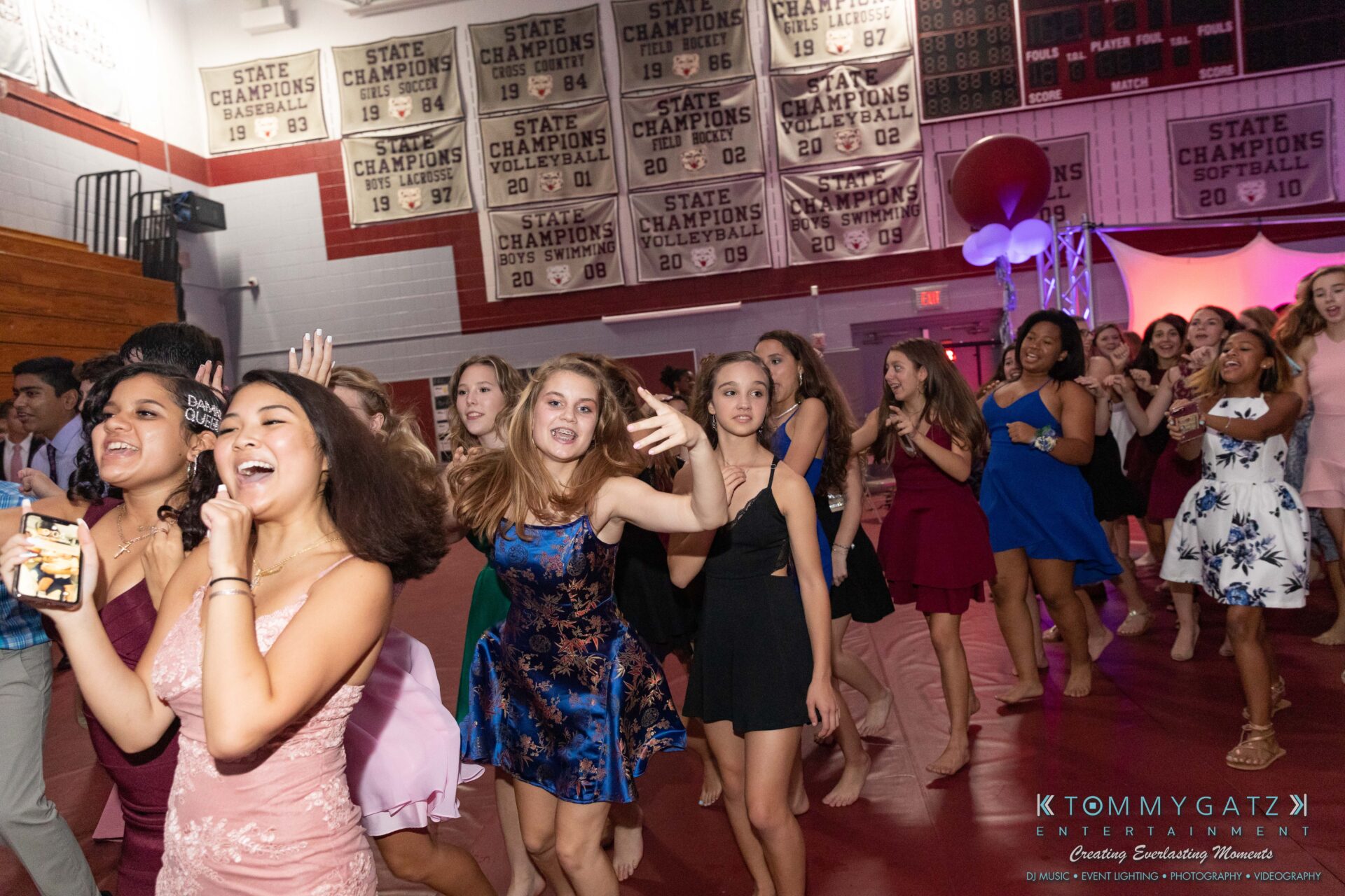 092819-Broadneck-Homecoming-Low-Res-WM-93 - TOMMY GATZ ENTERTAINMENT A group of young people dressed in formal attire dance energetically in a gymnasium, surrounded by championship banners.