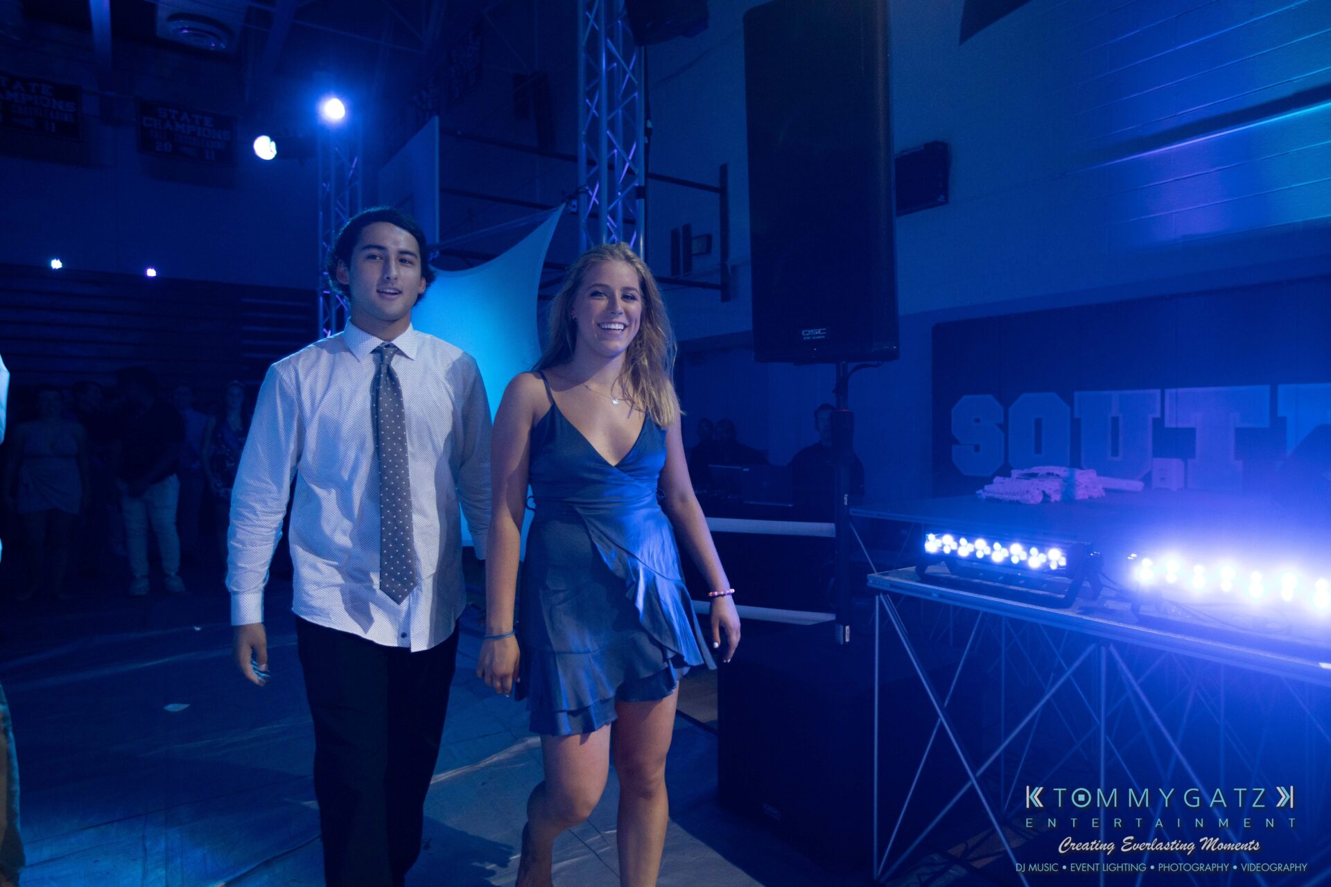100519-South-River-HC-Dance-Low-Res-WM-101 - TOMMY GATZ ENTERTAINMENT A young man and woman in formal attire walk on a dimly lit indoor dance floor with blue lighting.