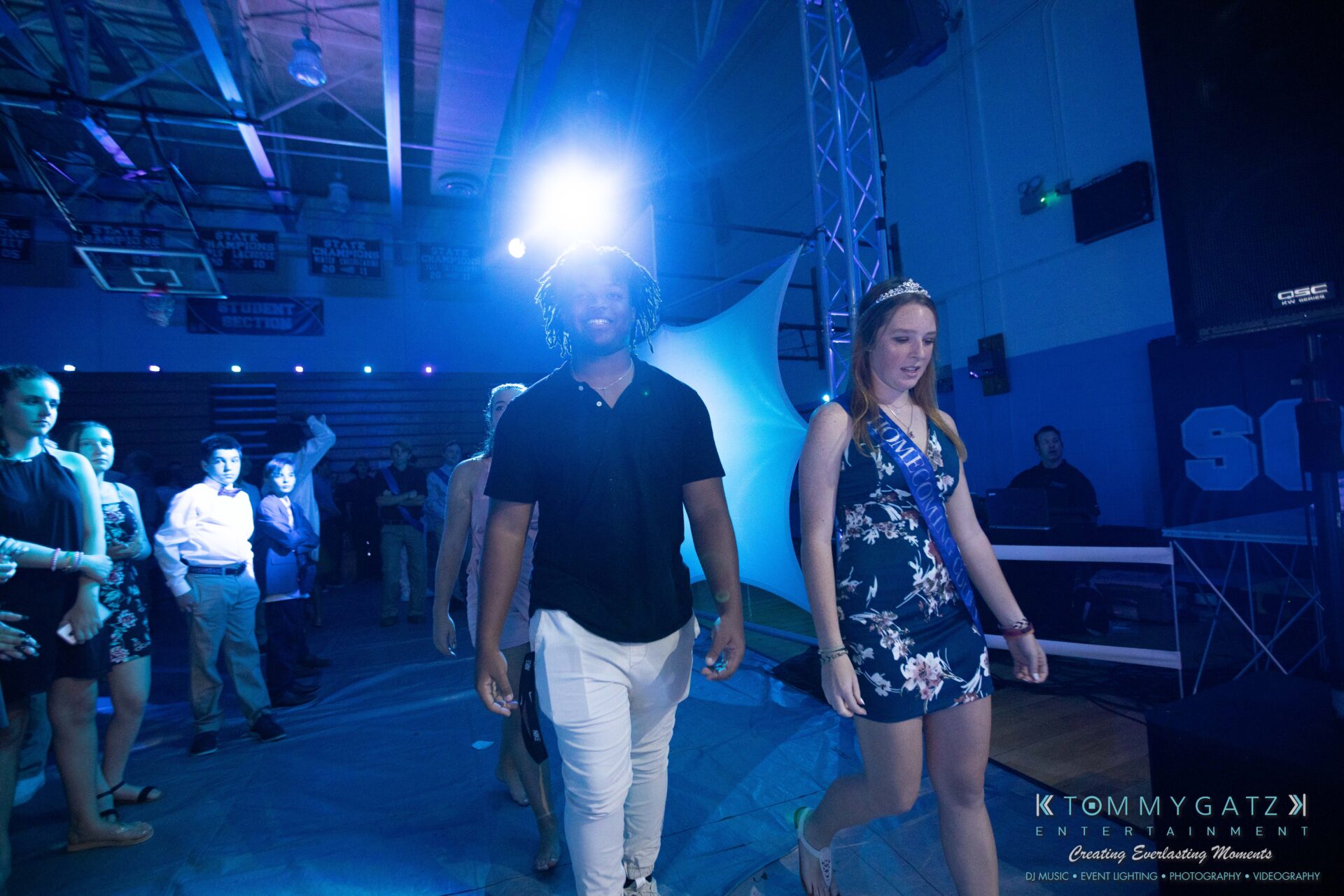 100519-South-River-HC-Dance-Low-Res-WM-102 - TOMMY GATZ ENTERTAINMENT People walk on a dimly lit stage with blue lighting, surrounded by a small audience. A woman wears a sash and a man is beside her.