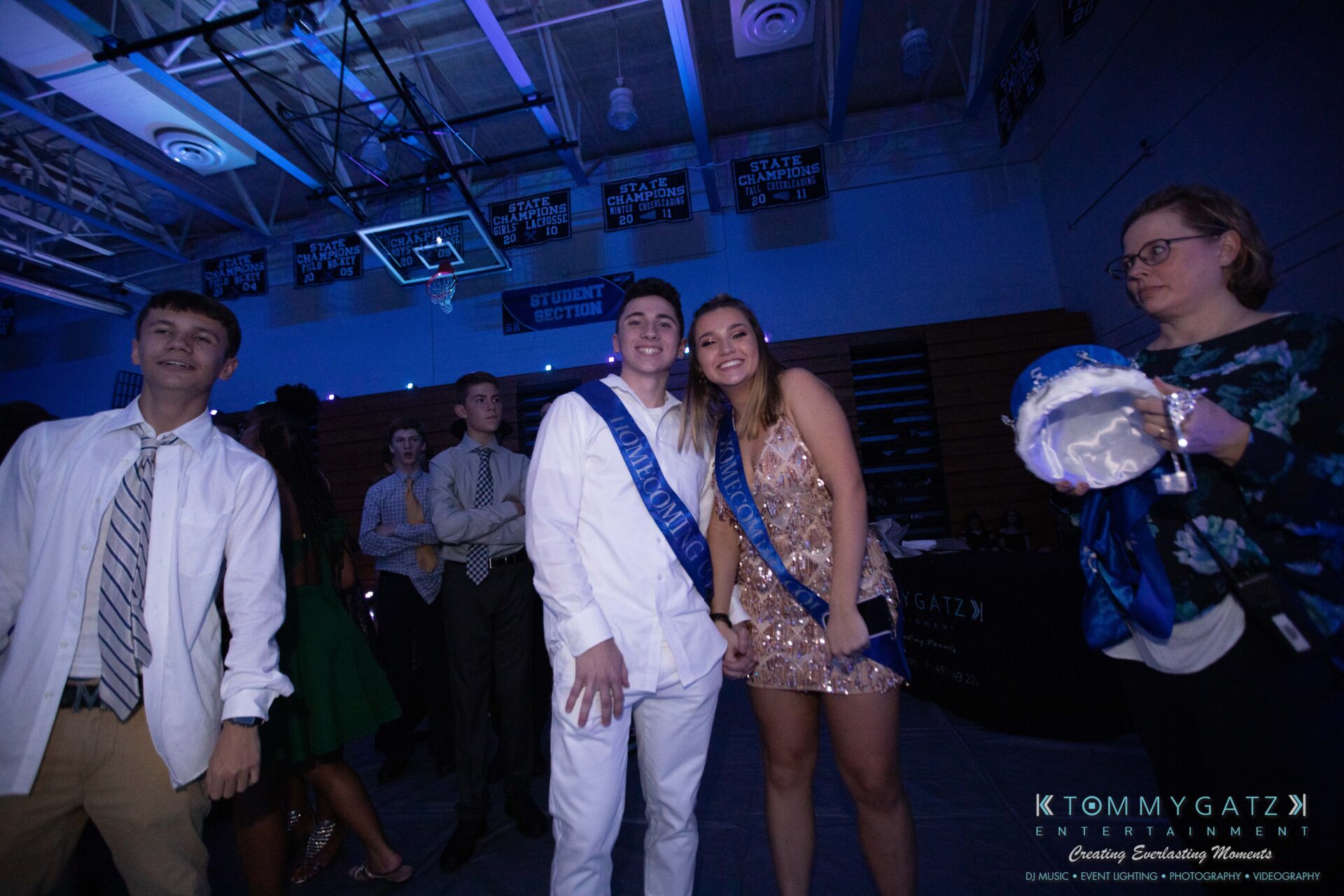 100519-South-River-HC-Dance-Low-Res-WM-103 - TOMMY GATZ ENTERTAINMENT Group of people in formal attire at a dimly lit indoor event. A couple in sashes stands at the center, smiling.