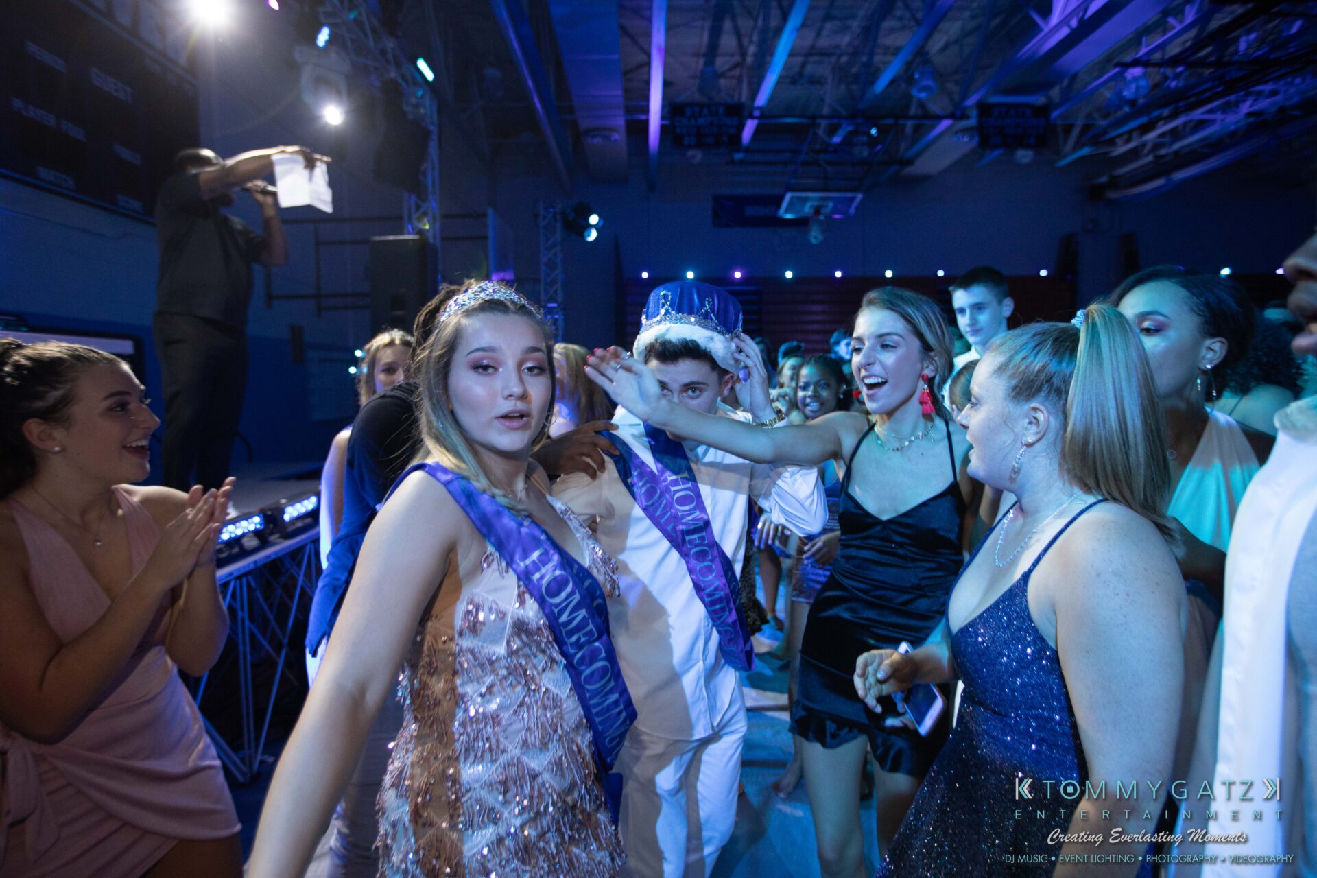 100519-South-River-HC-Dance-Low-Res-WM-105 - TOMMY GATZ ENTERTAINMENT A person places a crown on another person's head during an event with a crowd watching. They wear sashes and formal attire in a venue with blue lighting.