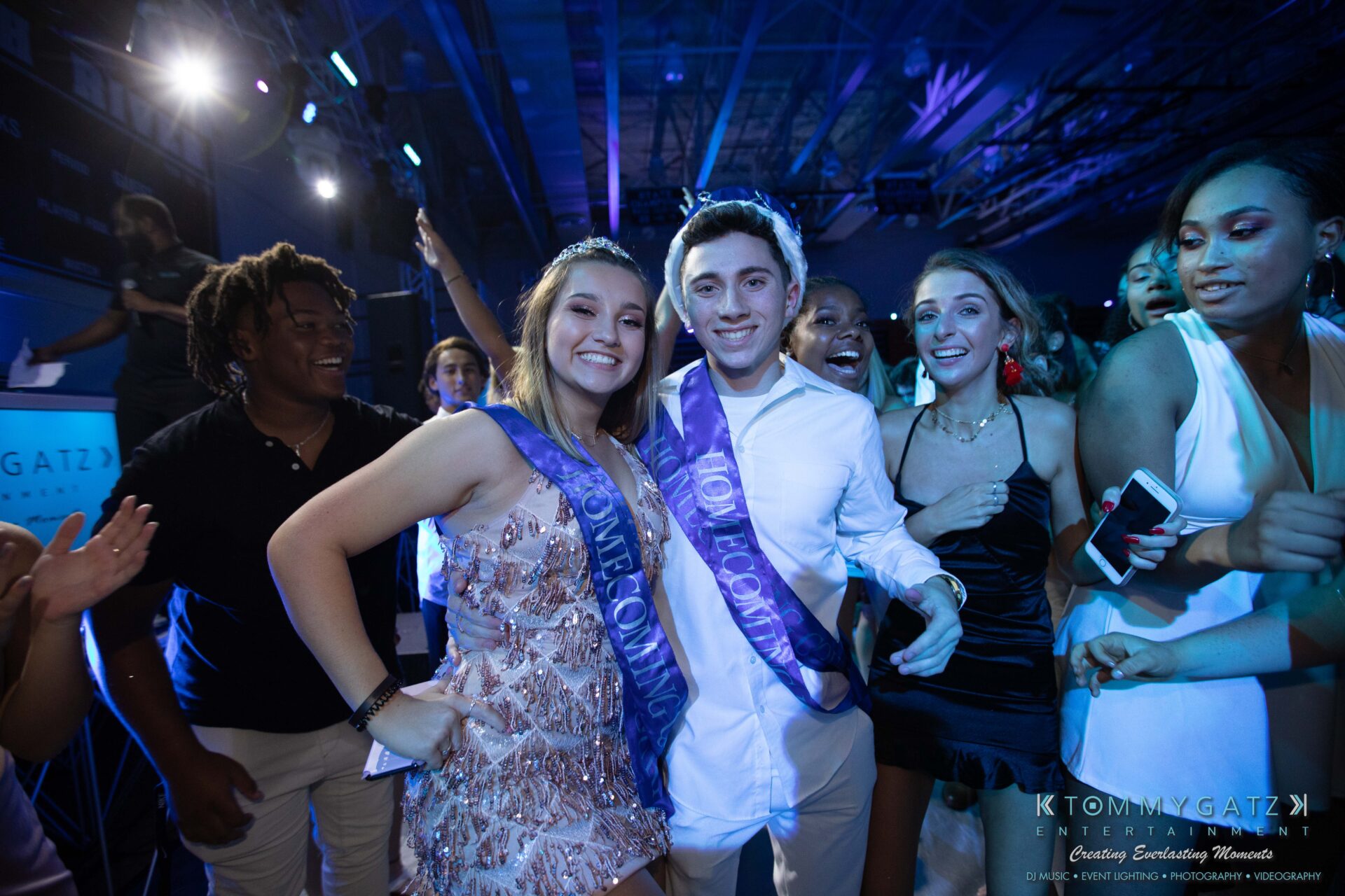 100519-South-River-HC-Dance-Low-Res-WM-107 - TOMMY GATZ ENTERTAINMENT Group of people, including two wearing "Homecoming 2023" sashes, smiling and posing in a lively party atmosphere with blue lighting.