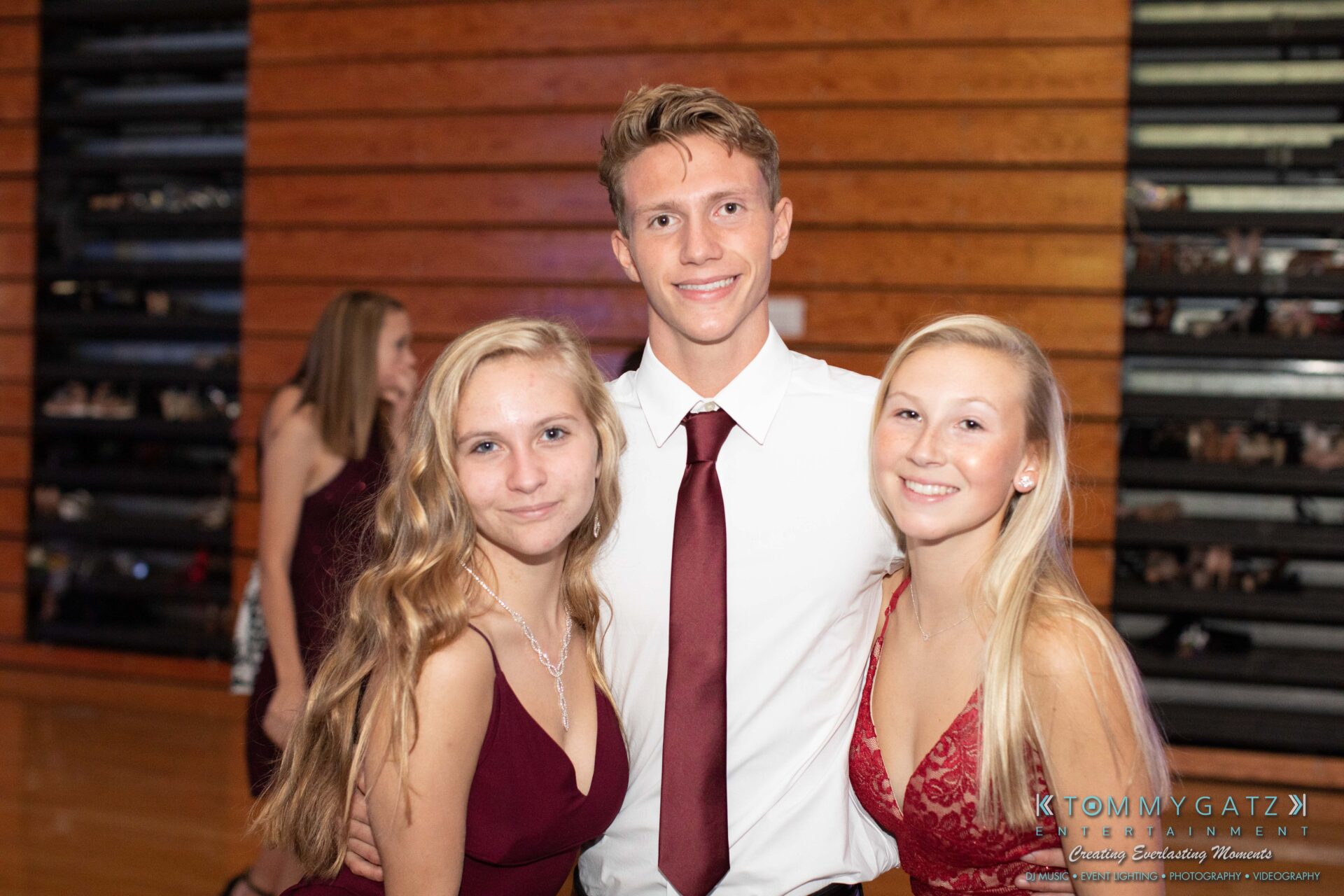 100519-South-River-HC-Dance-Low-Res-WM-15 - TOMMY GATZ ENTERTAINMENT Three people posing together, two women and a man, in a gym with wooden bleachers. They are dressed in formal attire.