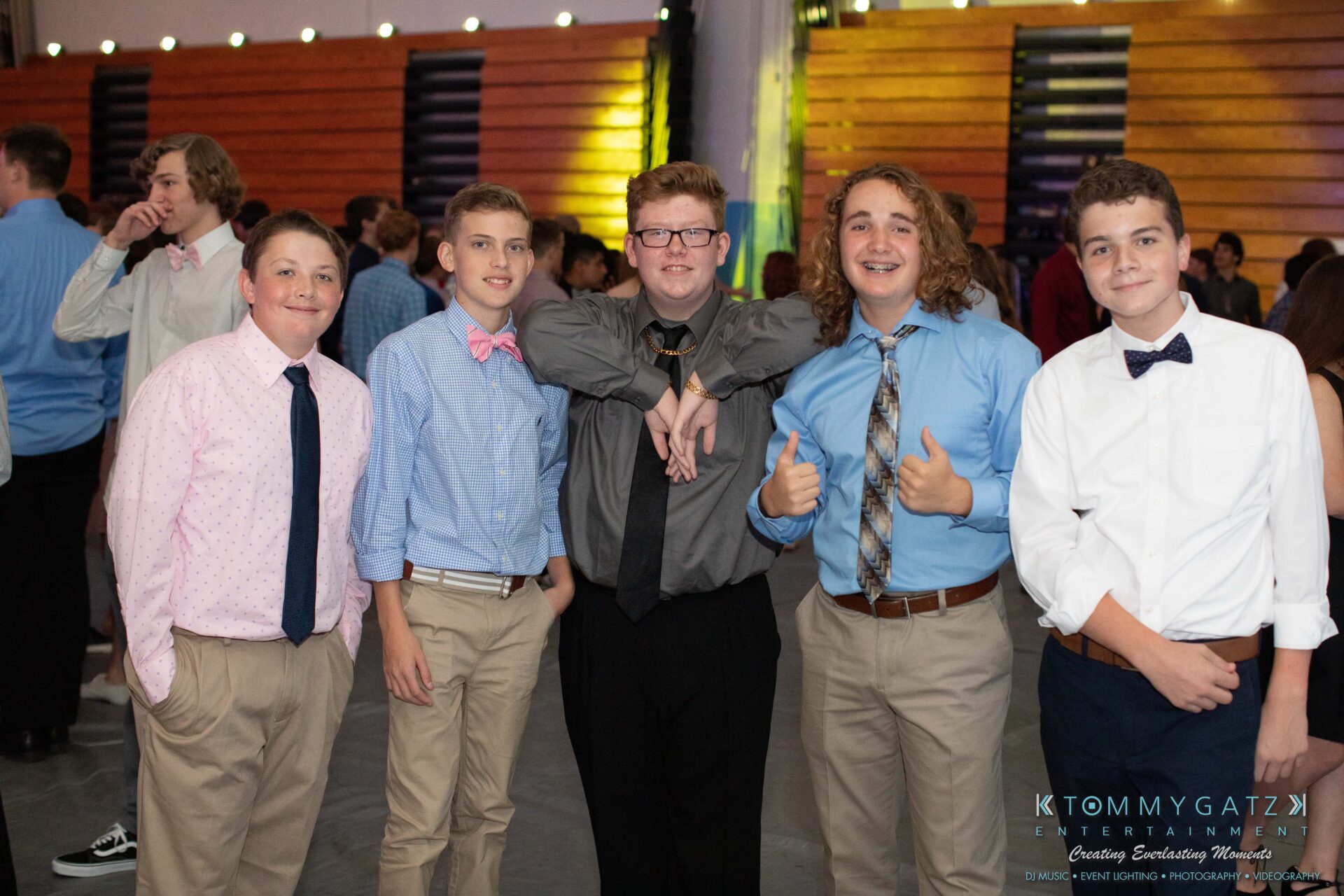 100519-South-River-HC-Dance-Low-Res-WM-23 - TOMMY GATZ ENTERTAINMENT Five young men dressed in formal attire pose together, some smiling and giving thumbs up, at a dance event in a hall with wooden walls and colorful lighting.