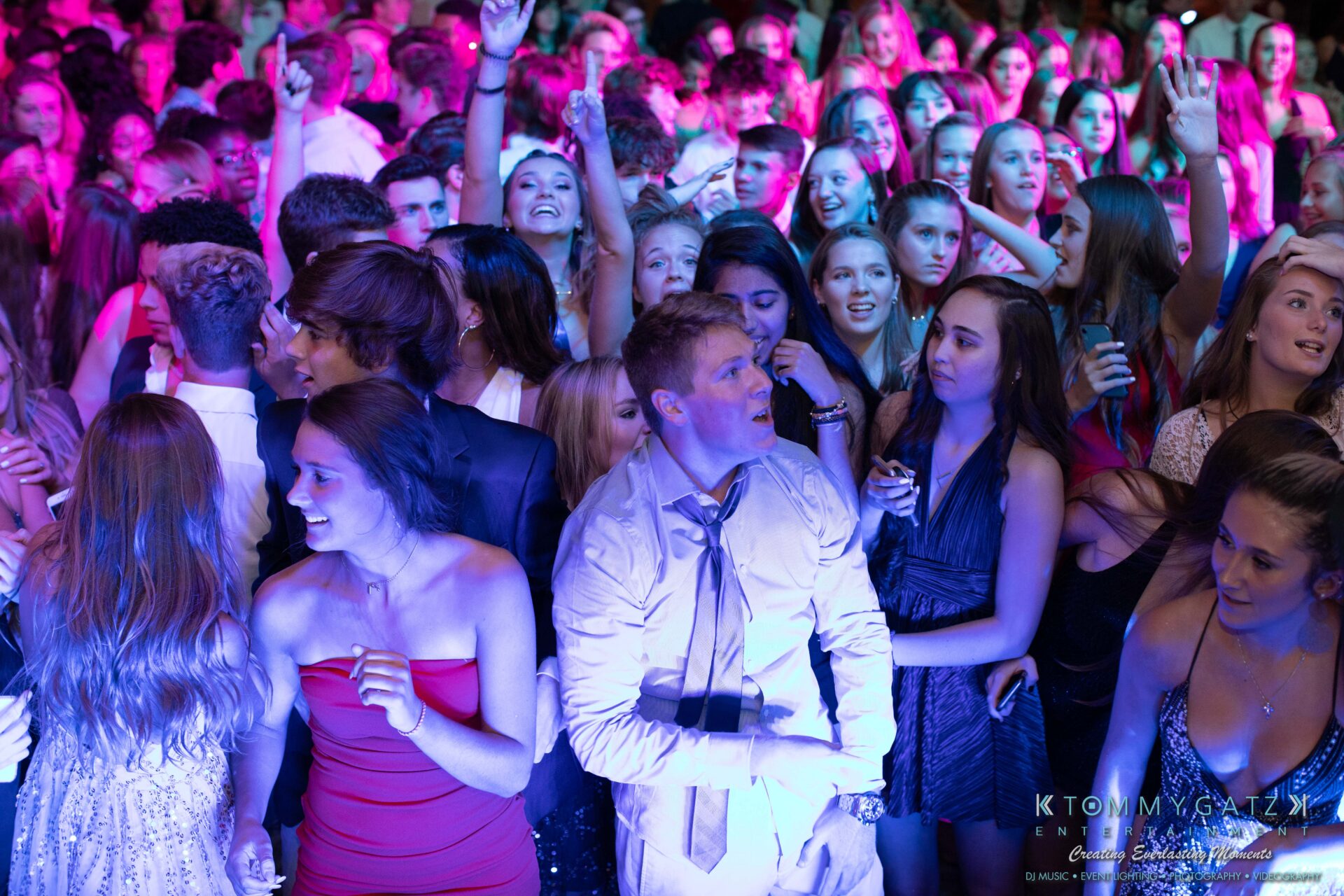 100519-South-River-HC-Dance-Low-Res-WM-27 - TOMMY GATZ ENTERTAINMENT A crowd of young people in formal attire dance and socialize under colorful lighting at an indoor event.