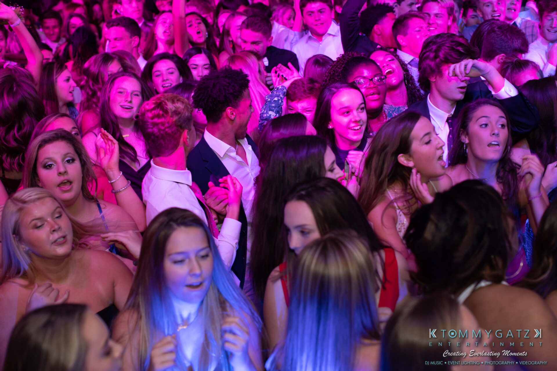 100519-South-River-HC-Dance-Low-Res-WM-32 - TOMMY GATZ ENTERTAINMENT A large group of people, dressed in formal attire, are gathered closely together at a lively dance event, bathed in pink and purple lights.