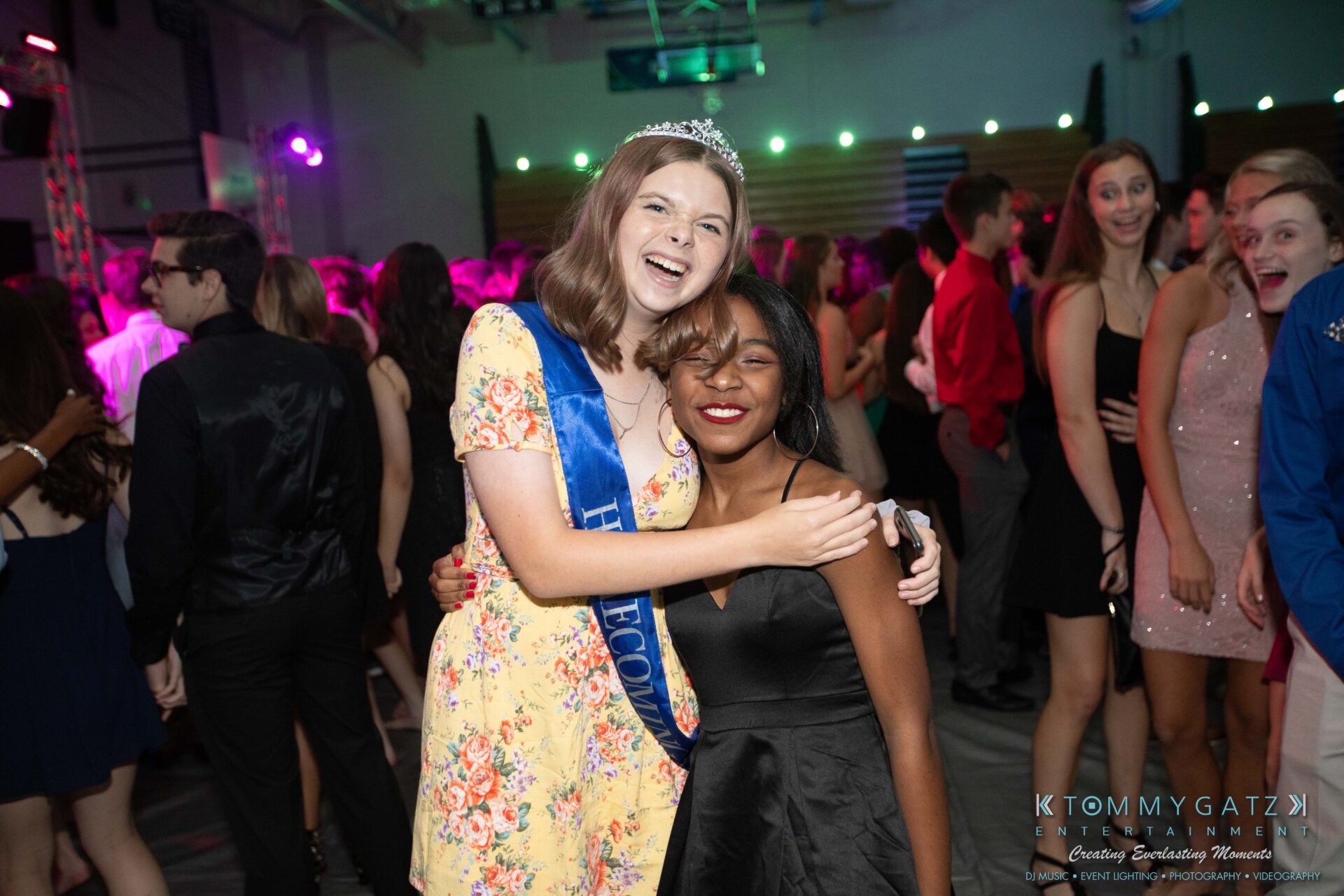 100519-South-River-HC-Dance-Low-Res-WM-36 - TOMMY GATZ ENTERTAINMENT Two young women, one wearing a "Homecoming" sash and crown, smile and embrace at a dance event. People socialize in the background.