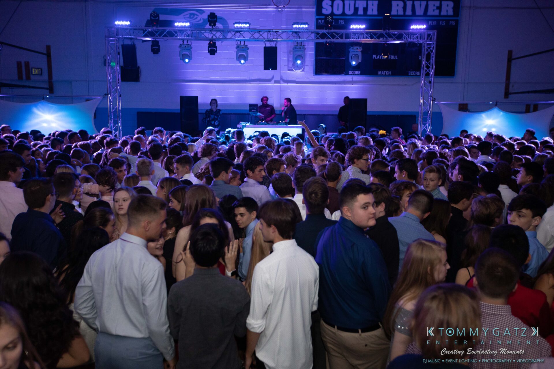 100519-South-River-HC-Dance-Low-Res-WM-68 - TOMMY GATZ ENTERTAINMENT A crowded school dance with students gathered on the dance floor. A DJ booth with two DJs and colorful lights are in the background.