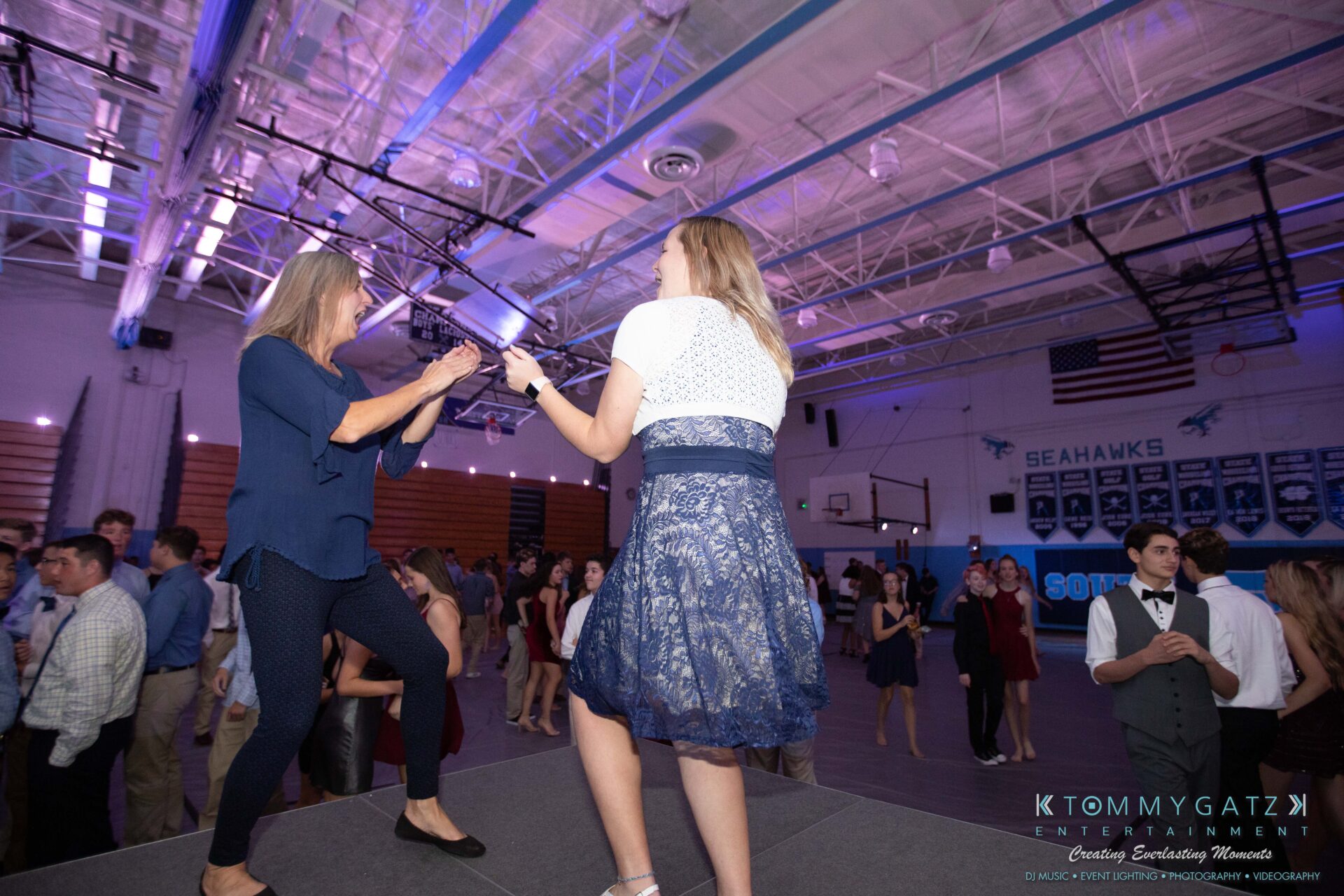 100519-South-River-HC-Dance-Low-Res-WM-69 - TOMMY GATZ ENTERTAINMENT Two women dance on a platform at a gymnasium event, surrounded by attendees in formal attire. Banners and overhead lighting are visible in the background.