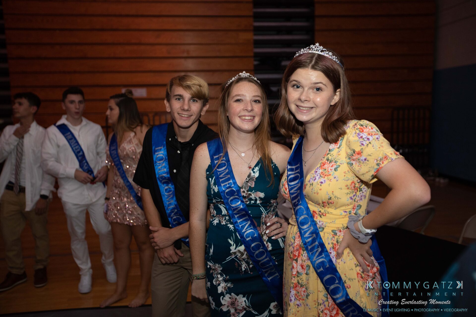 100519-South-River-HC-Dance-Low-Res-WM-84 - TOMMY GATZ ENTERTAINMENT Group of young people wearing "Homecoming Court" sashes stand indoors. Three individuals in the foreground wear tiaras and smile at the camera.