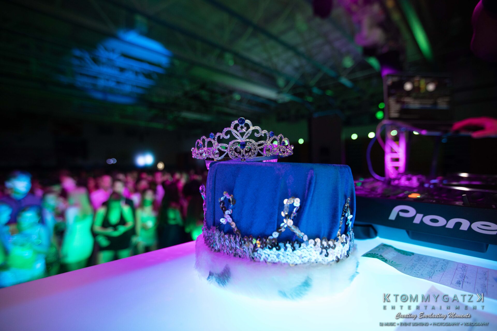 100519-South-River-HC-Dance-Low-Res-WM-85 - TOMMY GATZ ENTERTAINMENT A tiara on a blue velvet cushion sits on a table next to DJ equipment at a lively event with a crowd in the background.