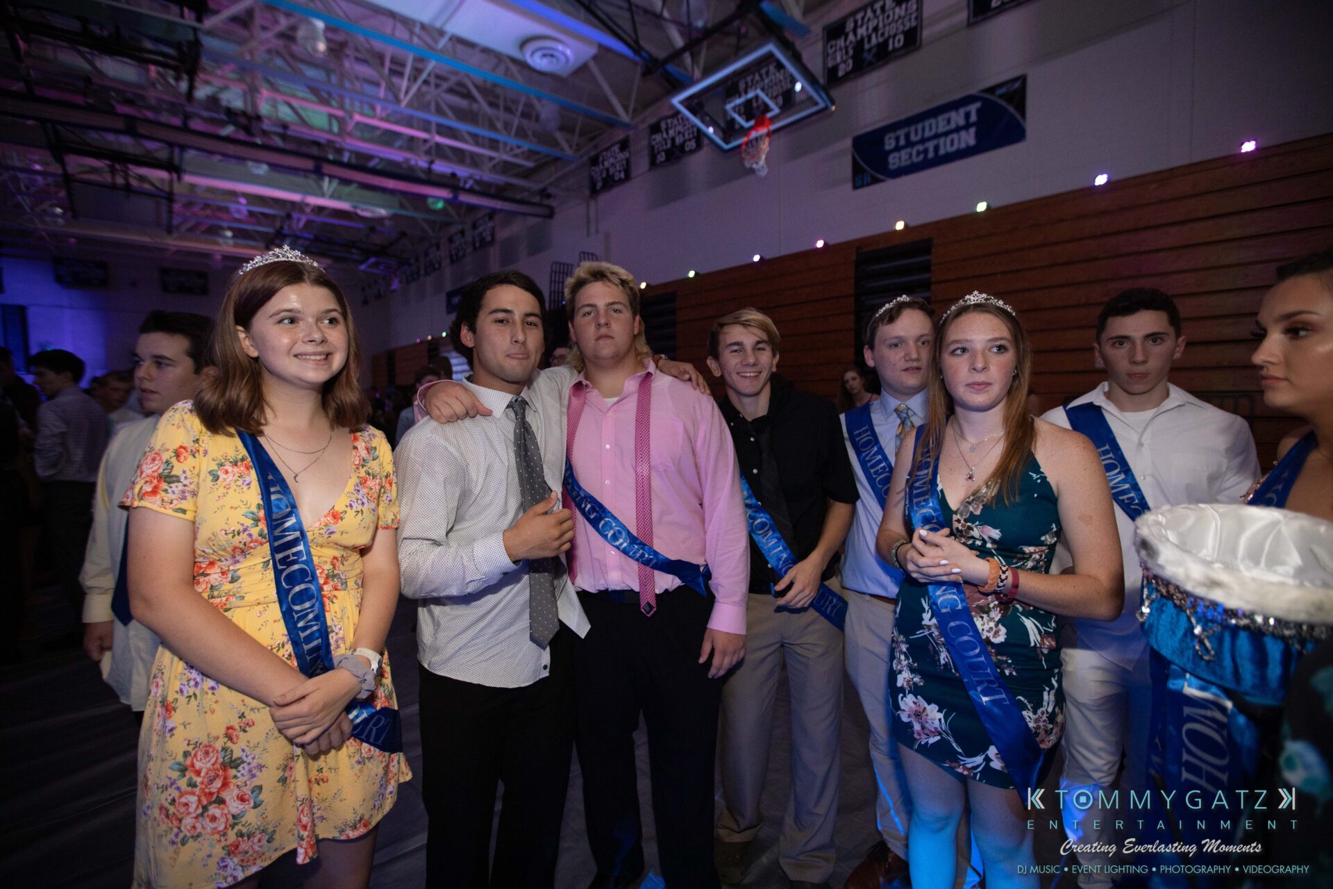 100519-South-River-HC-Dance-Low-Res-WM-87 - TOMMY GATZ ENTERTAINMENT A group of people wearing sashes and crowns stand together in a dimly lit room with a "Student Section" sign in the background.