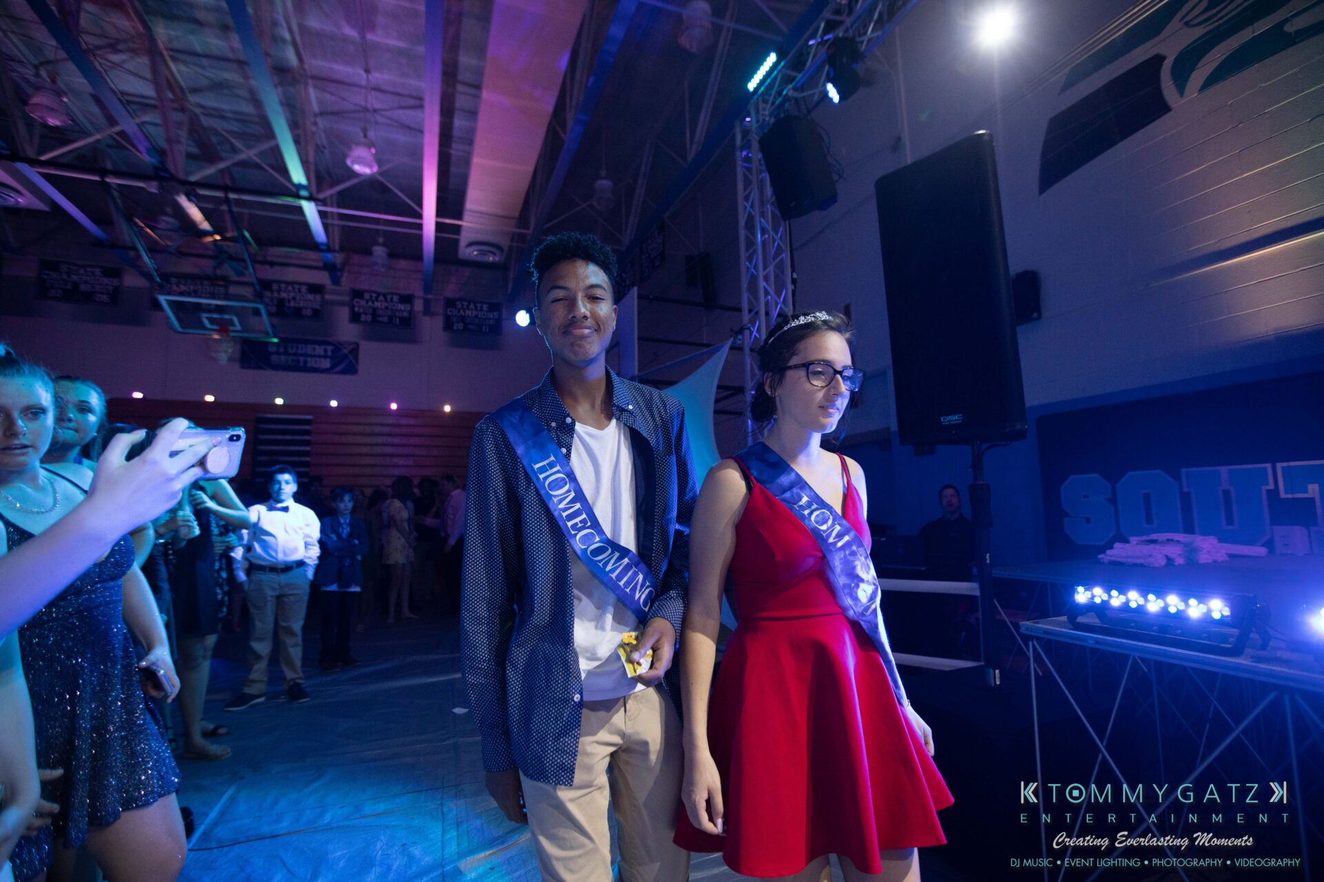 100519-South-River-HC-Dance-Low-Res-WM-97 - TOMMY GATZ ENTERTAINMENT A young man and woman, both wearing "Homecoming" sashes, walk together in a dimly lit venue with people in the background.