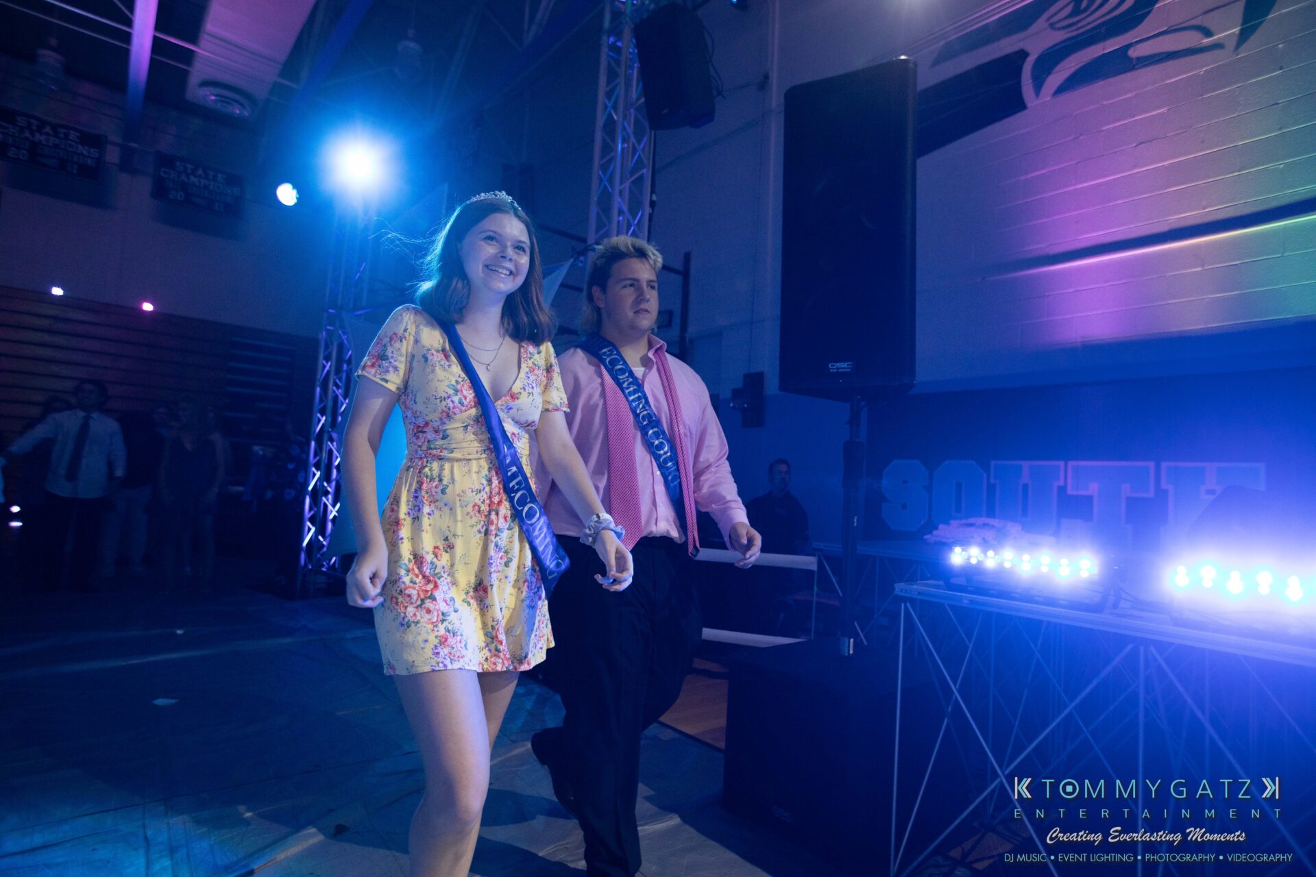 100519-South-River-HC-Dance-Low-Res-WM-99 - TOMMY GATZ ENTERTAINMENT A young woman and man, both wearing sashes, walk side by side in a dimly lit indoor venue.