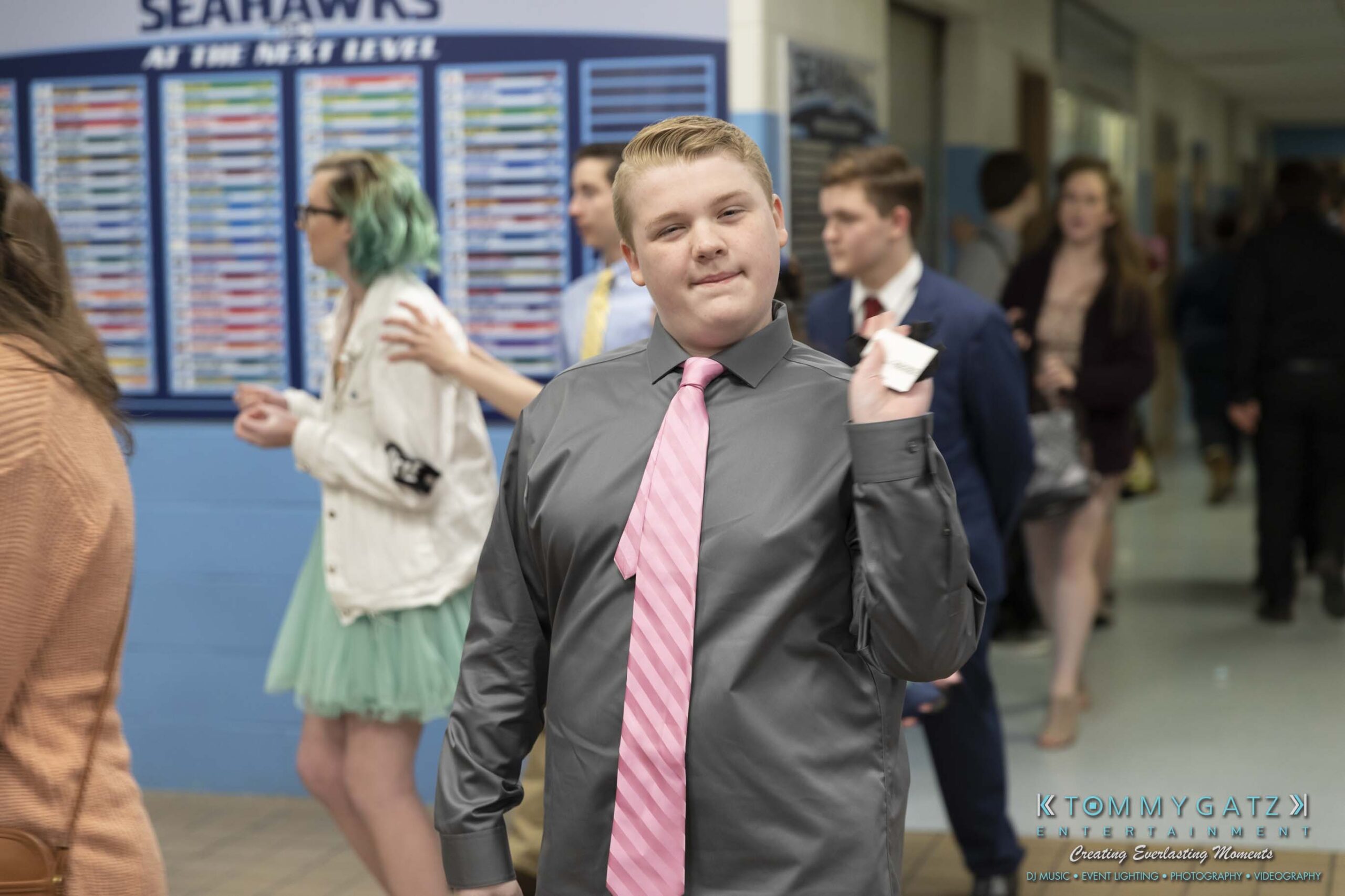SR-Spring-Fling-032319-FINALS-Low-Res-WM-16 - TOMMY GATZ ENTERTAINMENT Young person in a gray shirt and pink tie stands in a school hallway, holding a piece of paper. Other people are walking in the background.