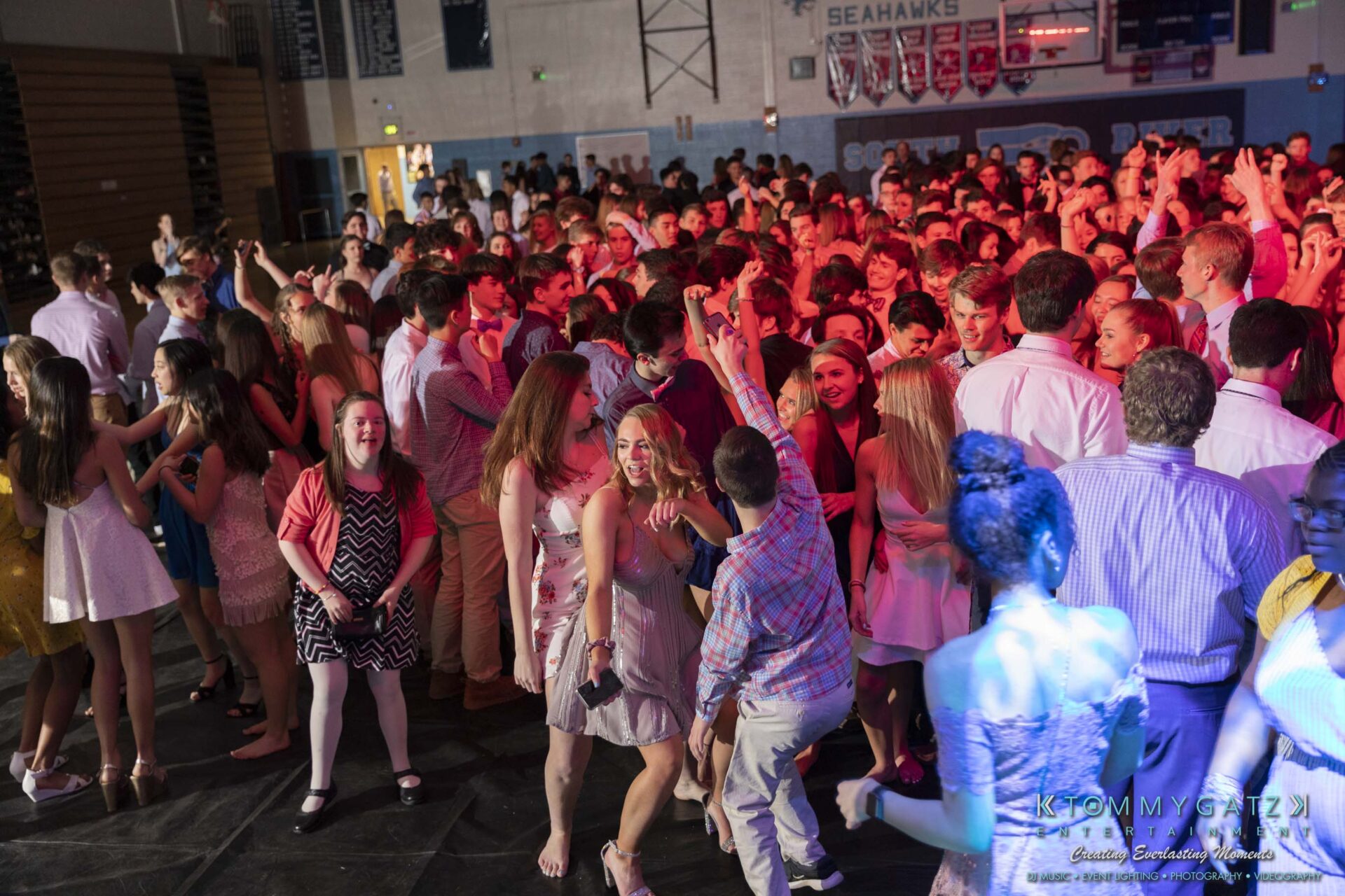SR-Spring-Fling-032319-FINALS-Low-Res-WM-30 - TOMMY GATZ ENTERTAINMENT A large group of people dancing and socializing at a gymnasium event, with colorful lighting and decorations in the background.