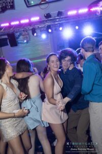 People dancing at a party under colorful lights. One person in a pink dress and another in a blue shirt are in focus.
