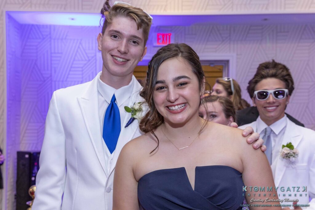A young woman in a dark blue dress and a young man in a white suit with a blue tie smile at the camera. Another person in sunglasses is in the background.