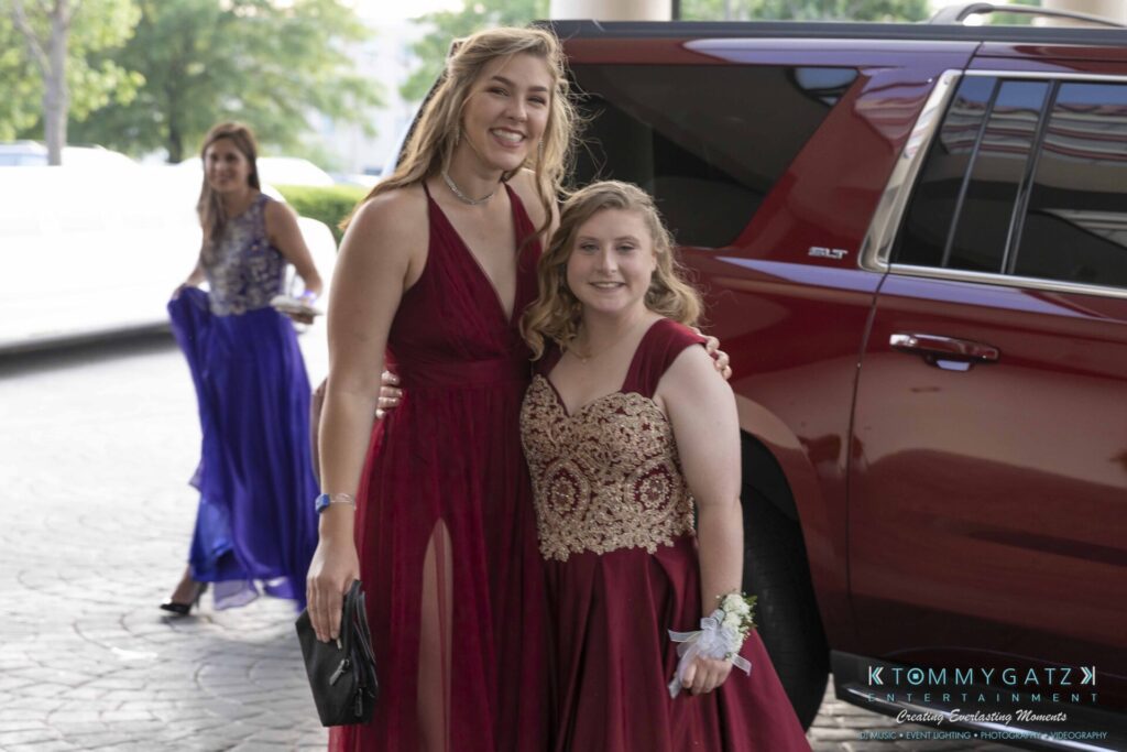 Two young women in formal dresses stand near a red vehicle, smiling at the camera. Another woman in a blue dress is in the background.