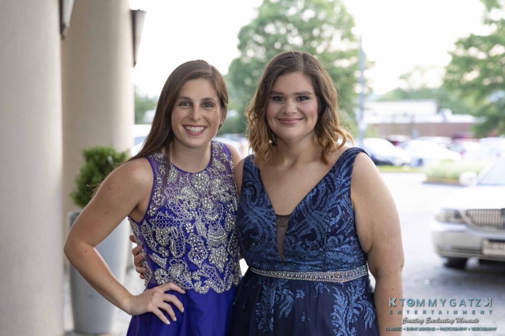 Two women in elegant blue dresses stand side by side, smiling, in an outdoor setting with cars and greenery in the background.