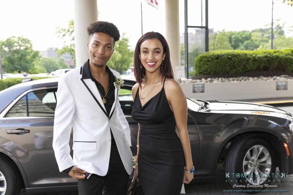 A man in a white suit and a woman in a black dress smile in front of a gray car outdoors.