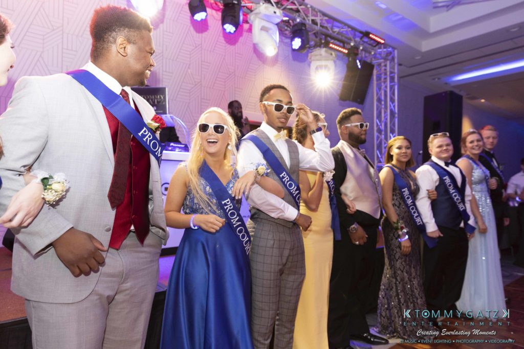 A group of people in formal attire and sashes stand on a stage, some wearing sunglasses. Stage lights are visible in the background.