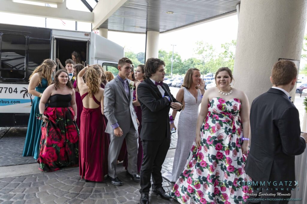 A group of people in formal attire wait in line outside a building near a bus.