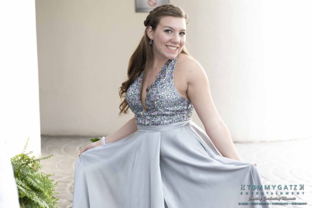 A woman in a sparkling silver gown poses playfully, lifting the skirt. She smiles brightly, standing on a light-colored floor with greenery nearby.