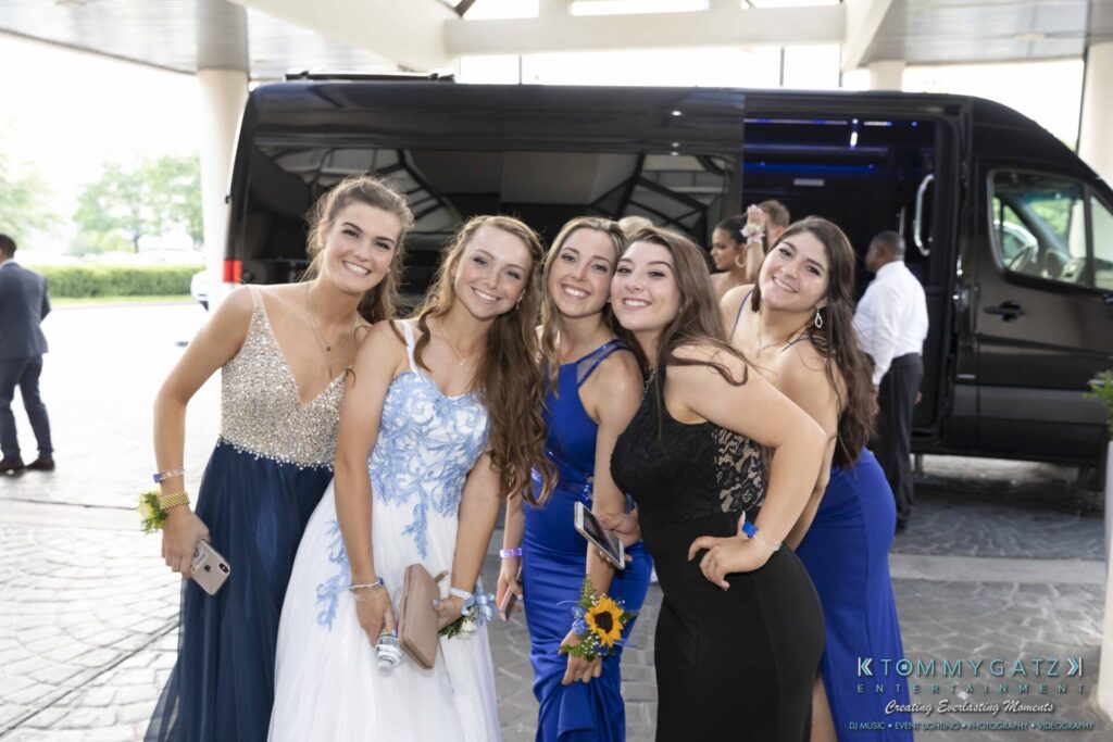 A group of five young women in formal dresses pose happily in front of a black vehicle in an outdoor setting.