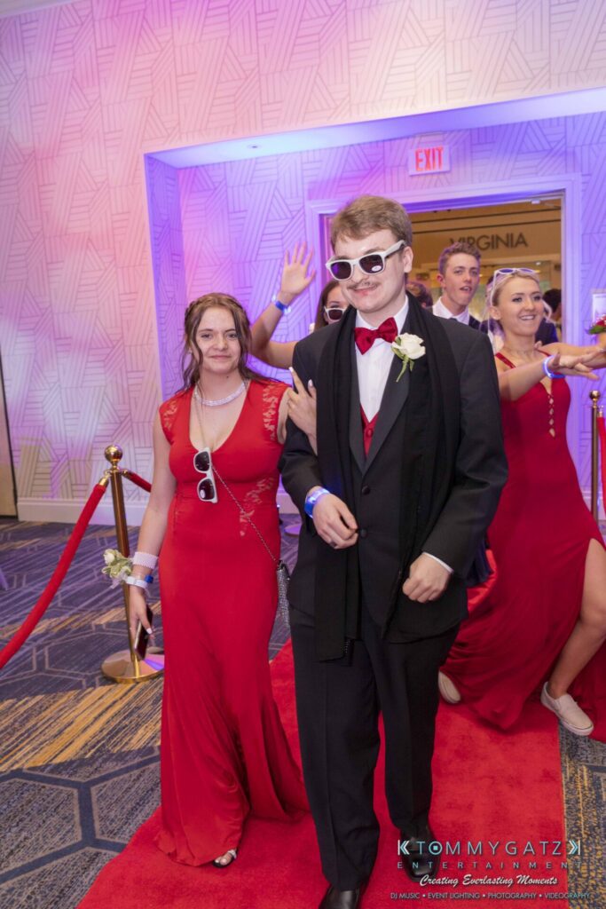 A young couple in formal attire walks on a red carpet. Both wear sunglasses and wristbands. Other people in similar outfits are visible in the background.